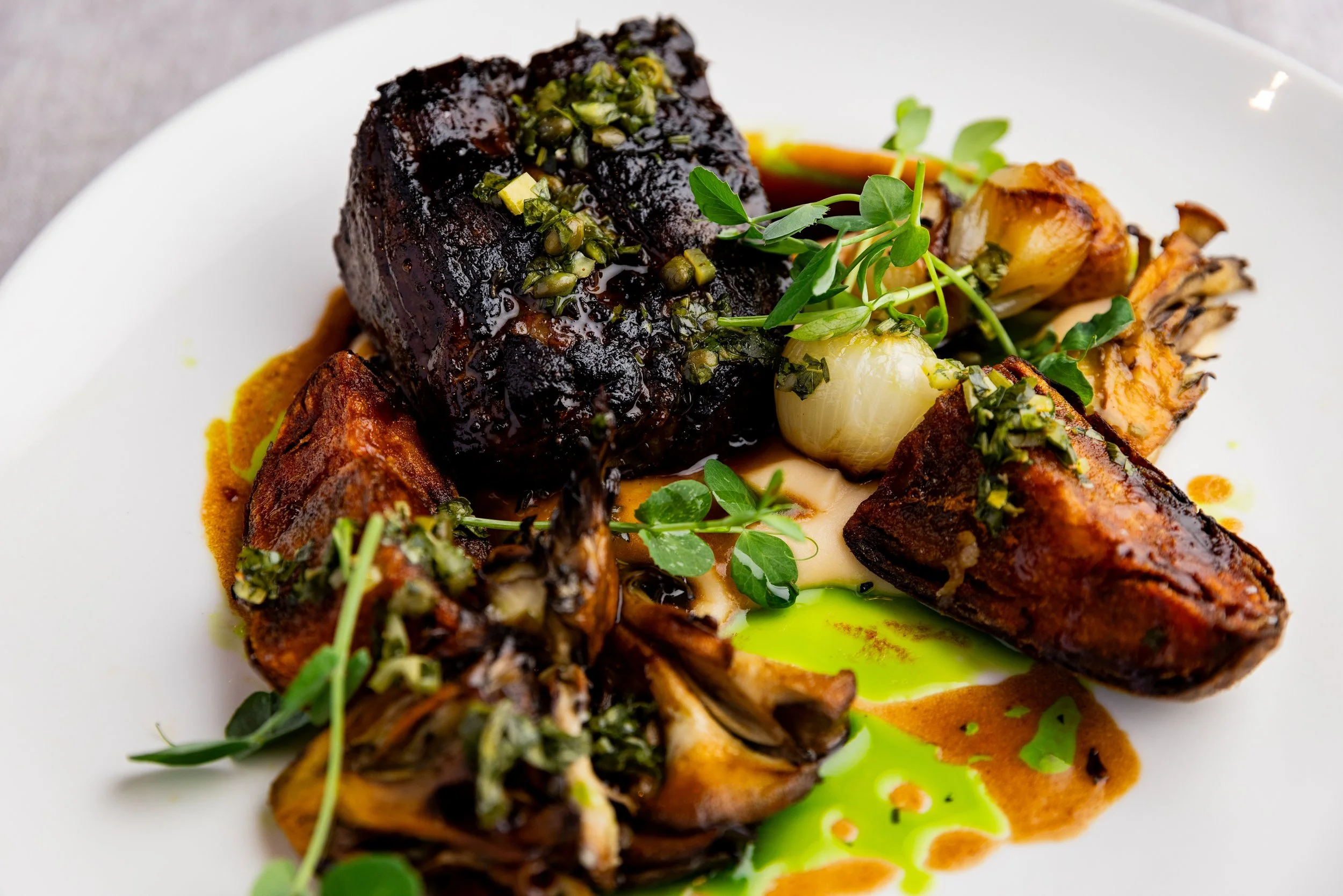 Close-up of a gourmet plate featuring a glazed, roasted meat, garnished with herbs, accompanied by roasted vegetables and colorful sauces on a white plate.