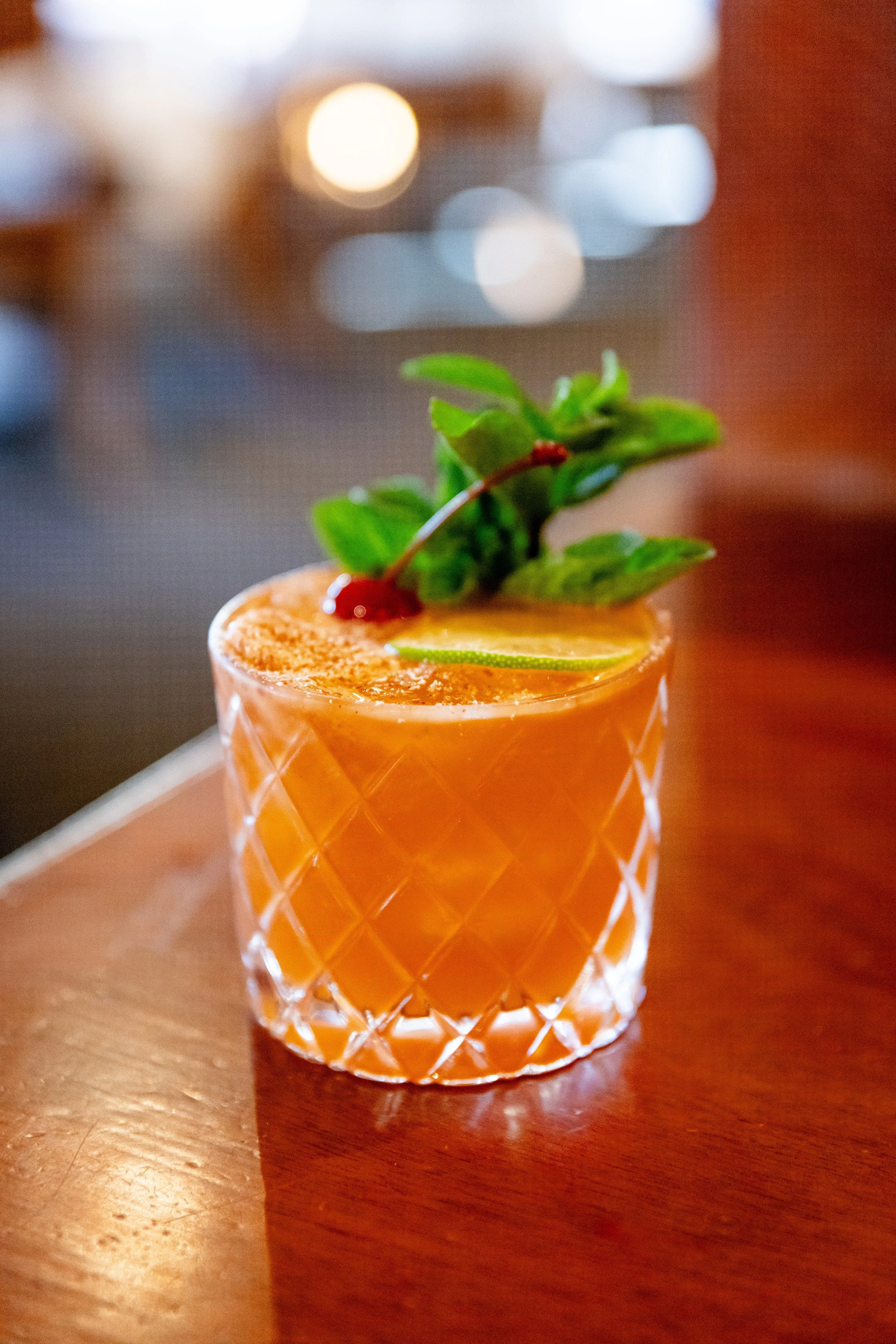 A cocktail garnished with a cherry, lime slice, mint leaves, and a sprinkle of cinnamon, served in a textured glass on a wooden table.