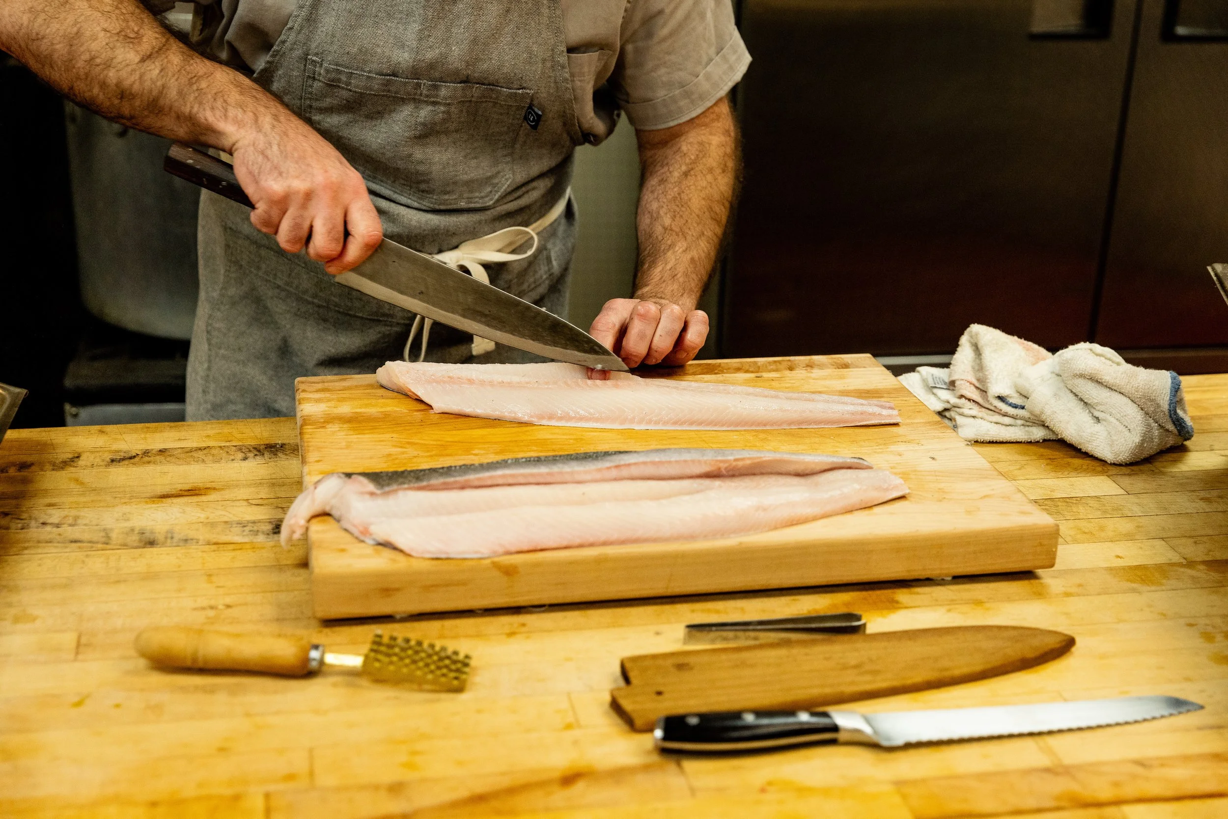 Person slicing raw fish fillets on a wooden cutting board in a kitchen.