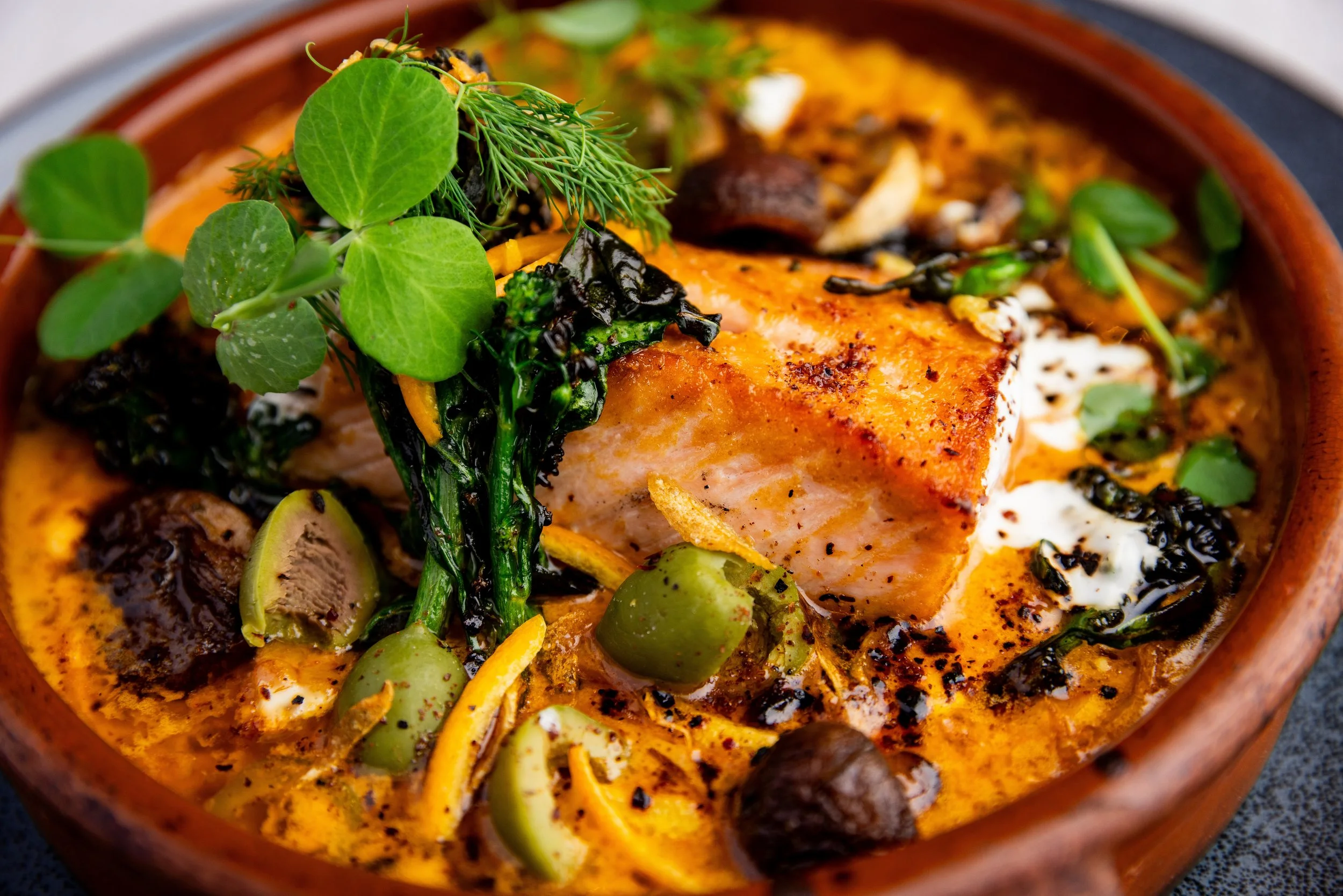 A bowl of salmon with vegetables and fresh herbs, served in a flavorful sauce.