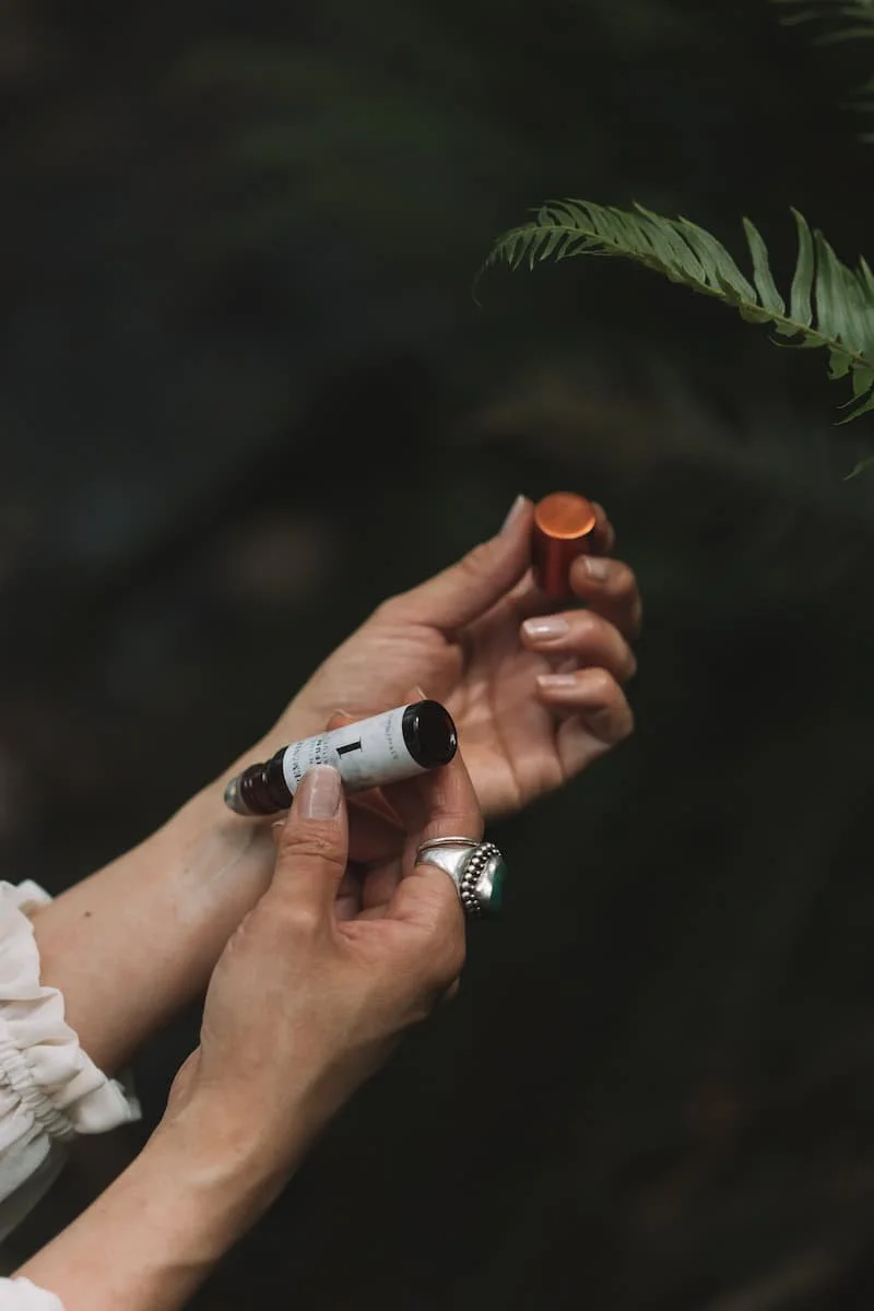 Person holding small brown glass bottle and a small copper container, with greenery in the background.