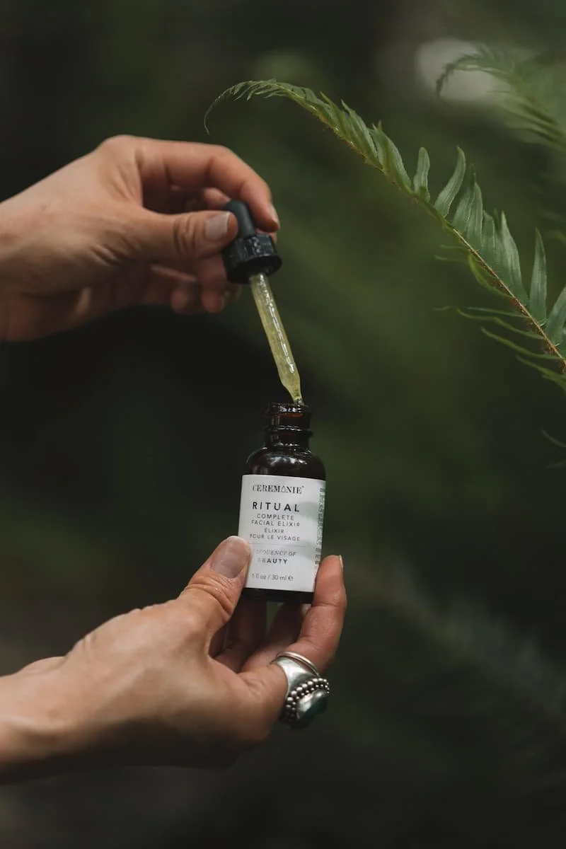 A person holding a bottle of Ritual Facial Elixir with a dropper, with a green fern leaf in the background.