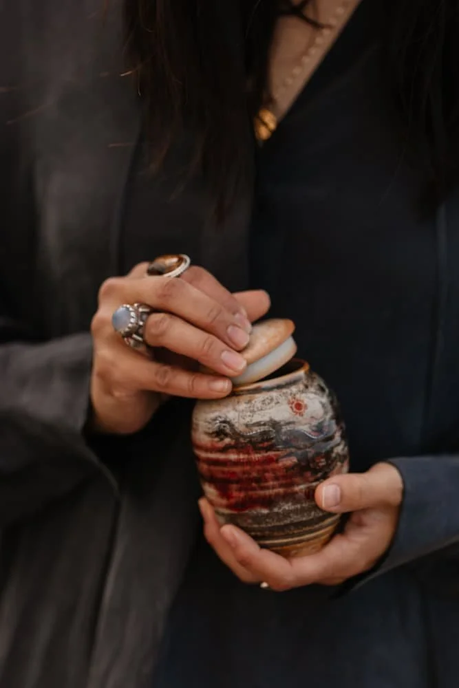 Person holding a small, ceramic, decorative container or vase with a red, black, and beige pattern, placing or removing a thin lid.
