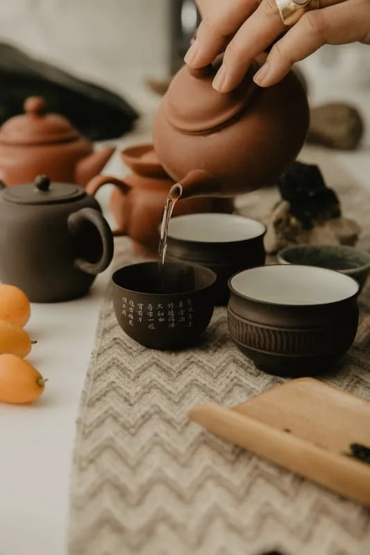 Person pouring tea from a clay teapot into a teacup, surrounded by various traditional teapots and bowls on a table with a patterned cloth.
