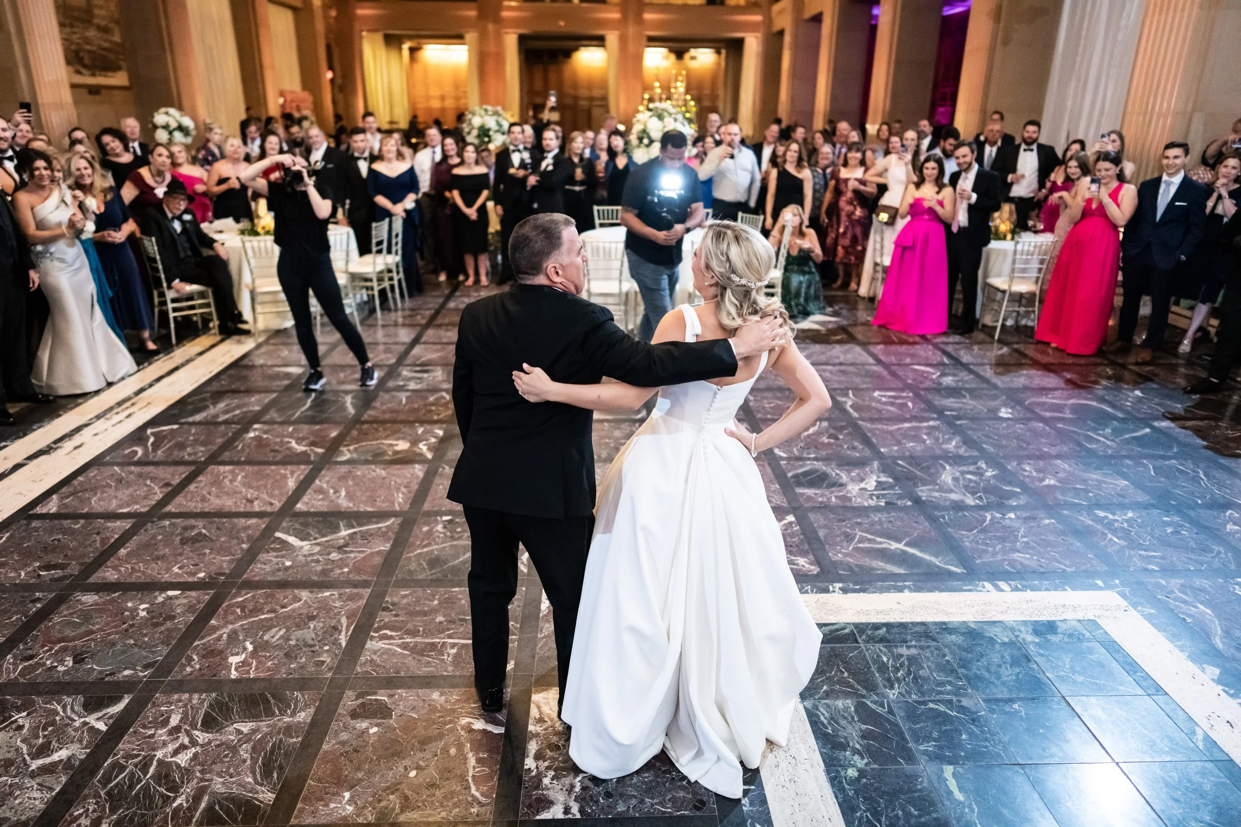 A man and a woman in formal attire dancing at a wedding reception with guests watching and a photographer capturing the moment in a large hall with elegant decor.