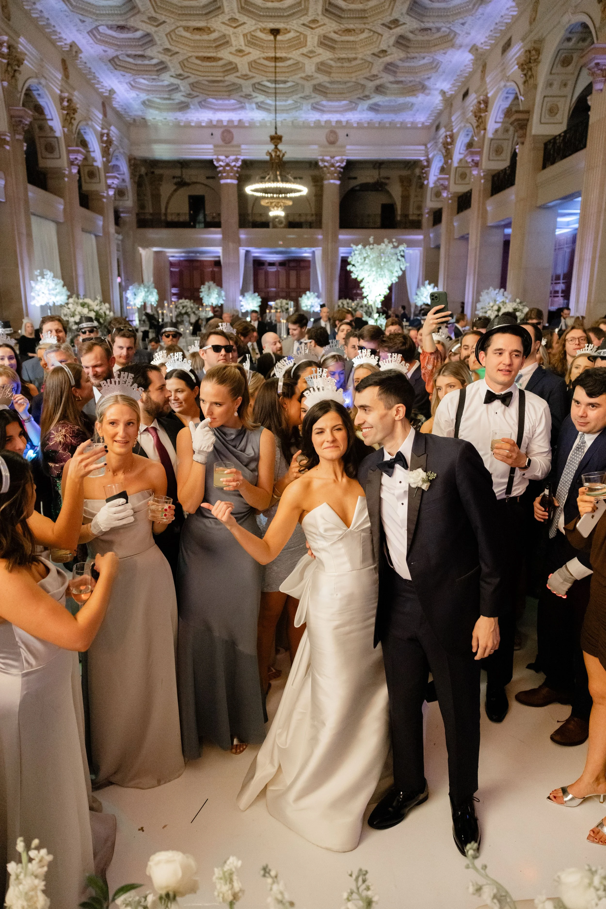 A large group of people celebrating at a wedding reception in an elegant venue with ornate ceilings. The bride in a white gown and groom in a tuxedo are at the center, surrounded by guests holding drinks and wearing celebratory accessories. The atmosphere is festive and lively.