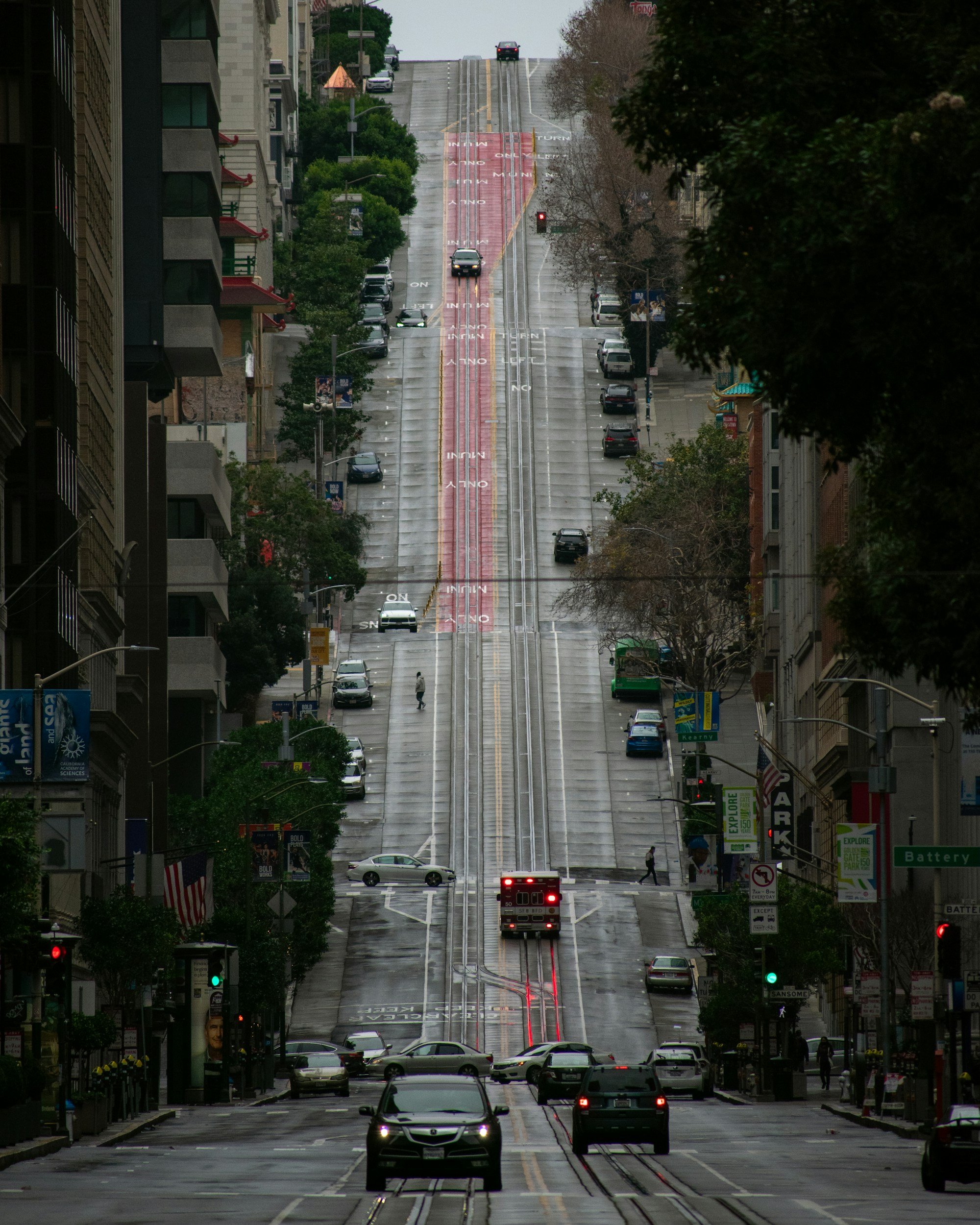 City street view with tram tracks running down the center, cars parked along both sides, traffic lights, and pedestrians, with a hill in the background and buildings lining the street.