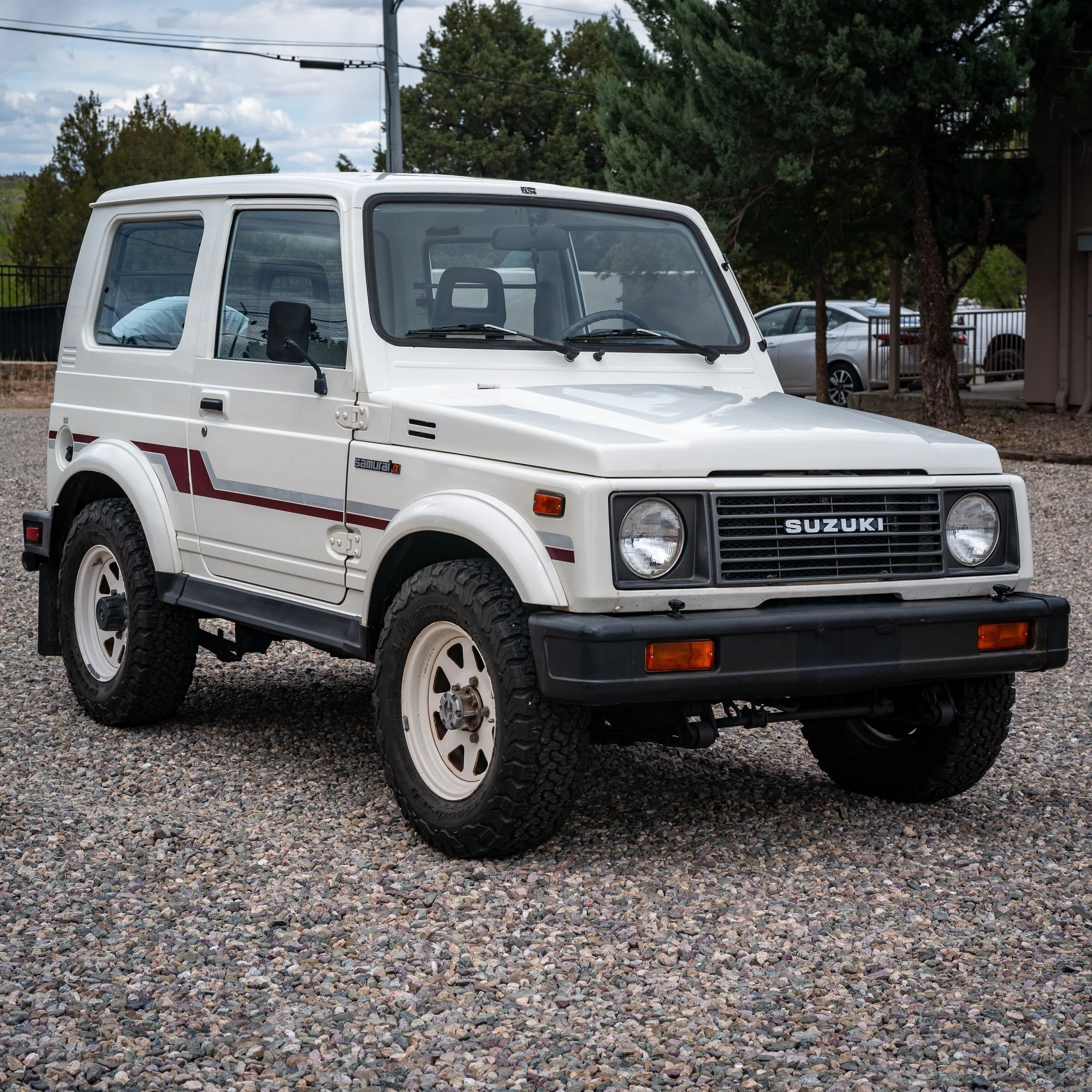 A white Suzuki Samurai SUV parked on a gravel surface with trees and parked cars in the background.