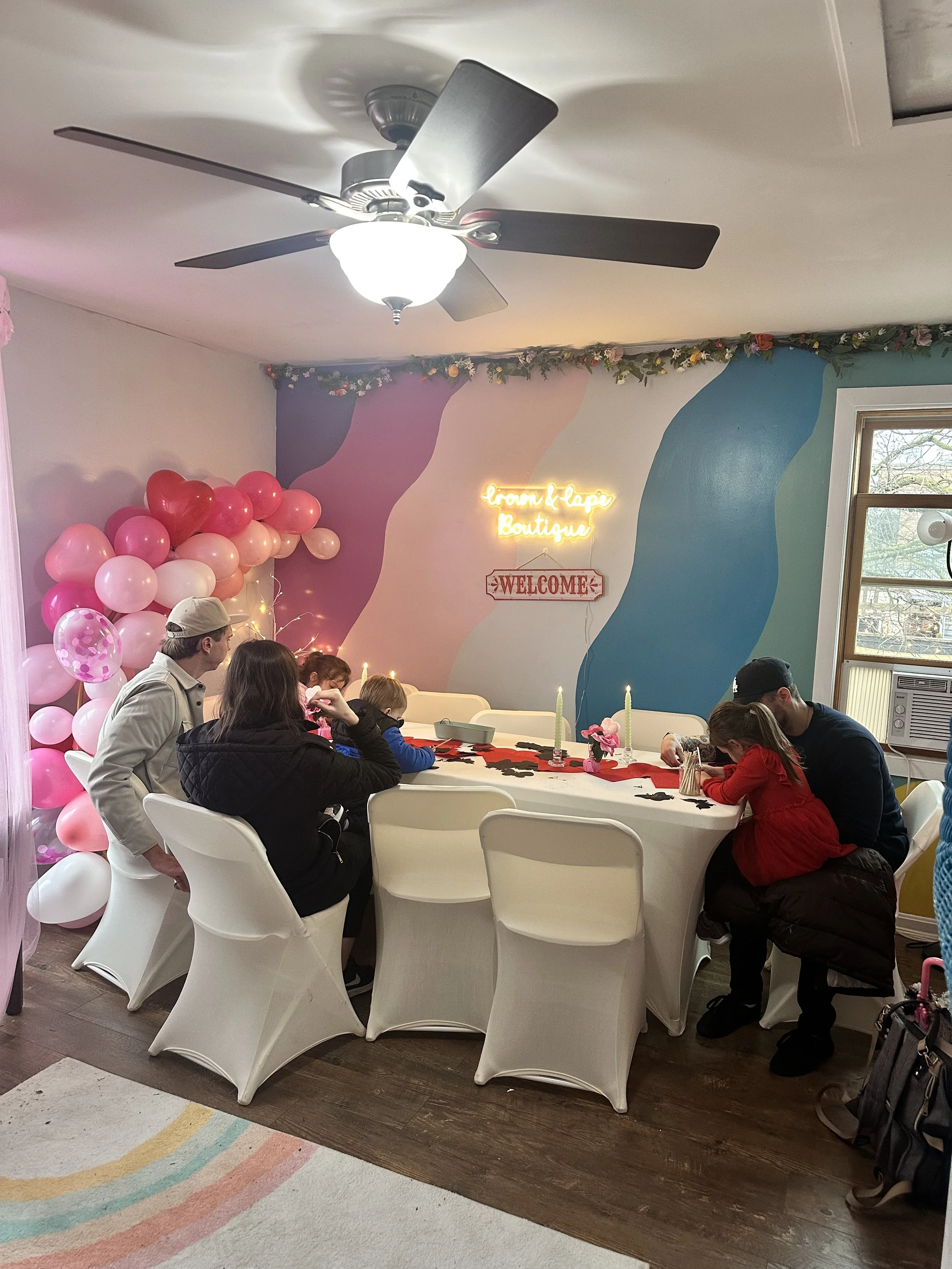 Children and adults gathered around a table decorated with pink candles and black star-shaped confetti in a boutique celebration space. A pink and red balloon arch is visible on the left, and colorful wall art with neon signage reading 'from & lave Boutique' and a welcome sign can be seen on the back wall. The room has a ceiling fan, a window, and festive decor.