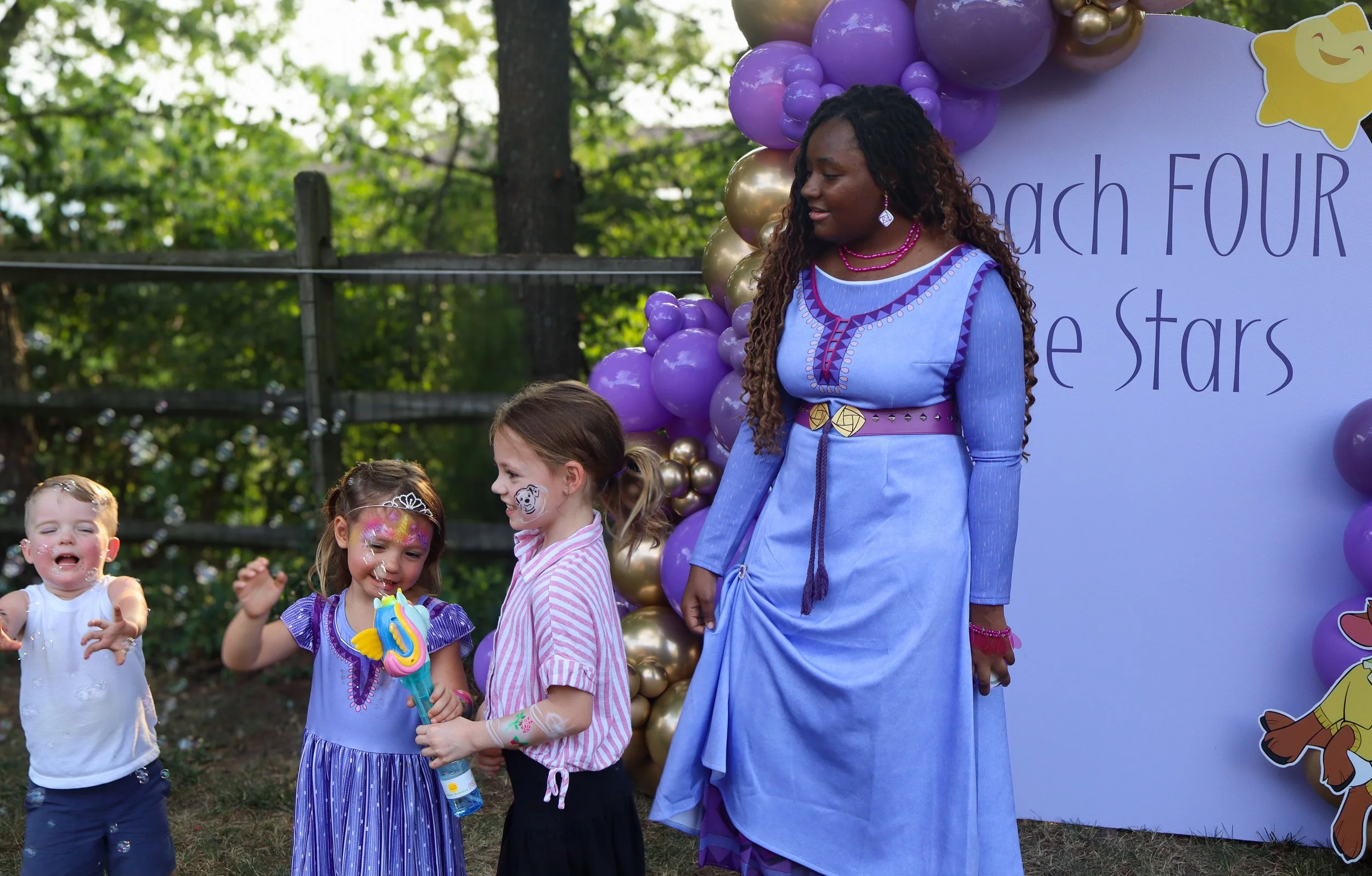 A woman dressed as a character from the animated series 'Inside Out' with four children outdoors at a celebration. The woman is wearing a blue dress with purple and gold accents, standing beside a decorated backdrop with balloons and cartoon characters. The children are happily interacting, with one holding a bubble gun and face paint visible on some children.