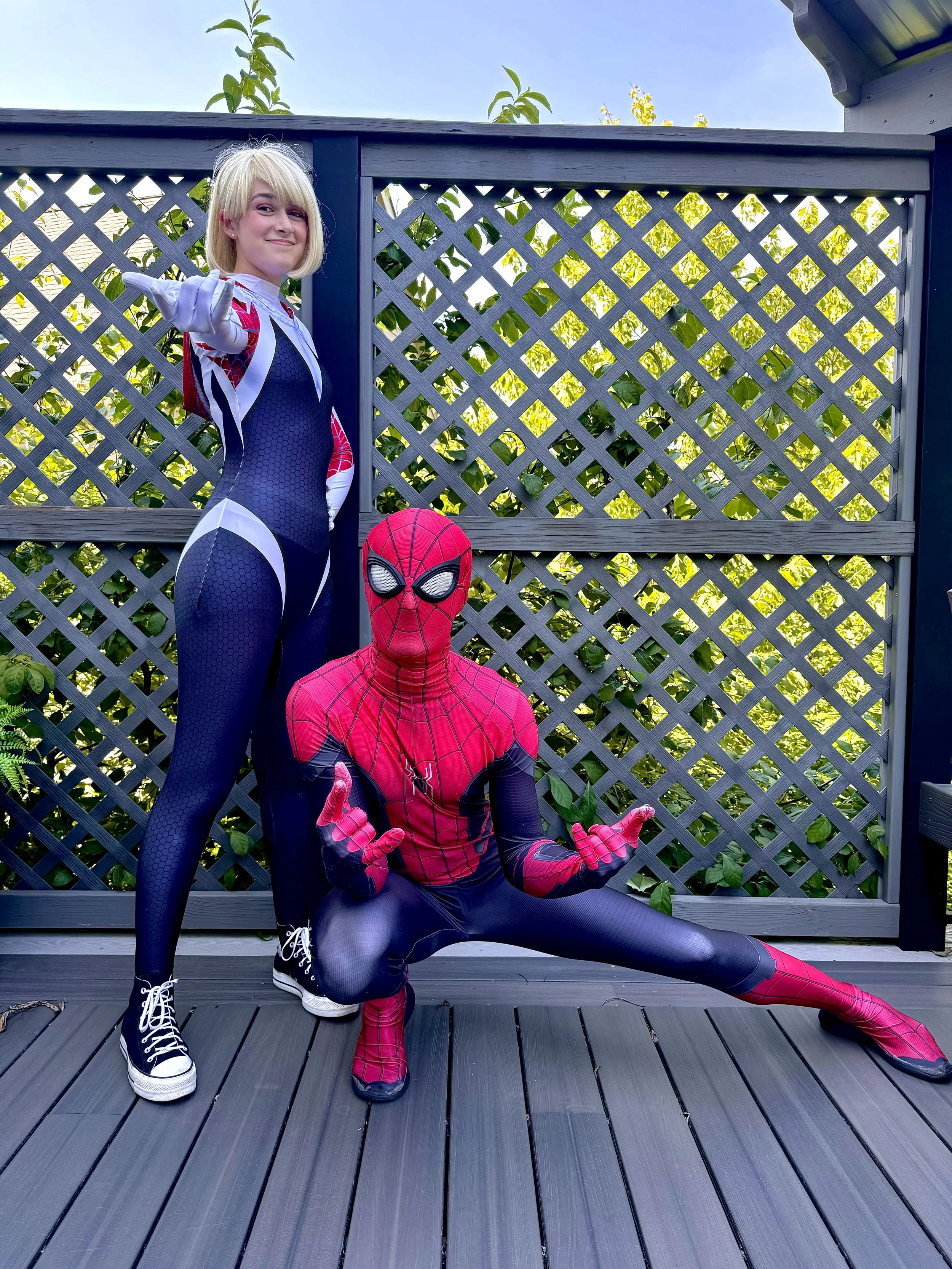 Two people dressed in Spider-Man and Spider-Girl costumes pose on a wooden deck in front of a fence with green plants behind them.