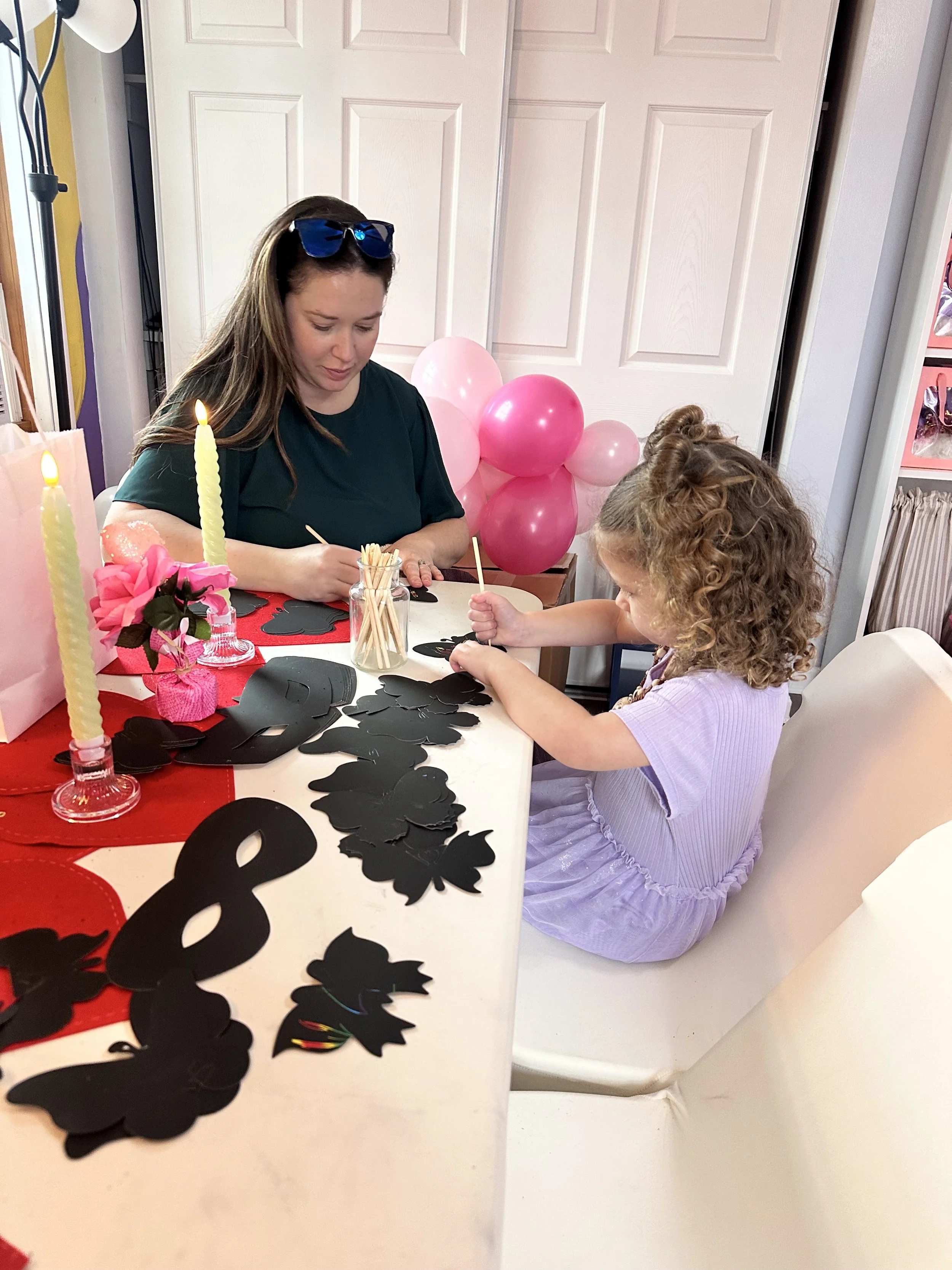 A woman and a young girl sitting at a table, decorating for a birthday party with black paper cutouts, pink balloons, and candles.