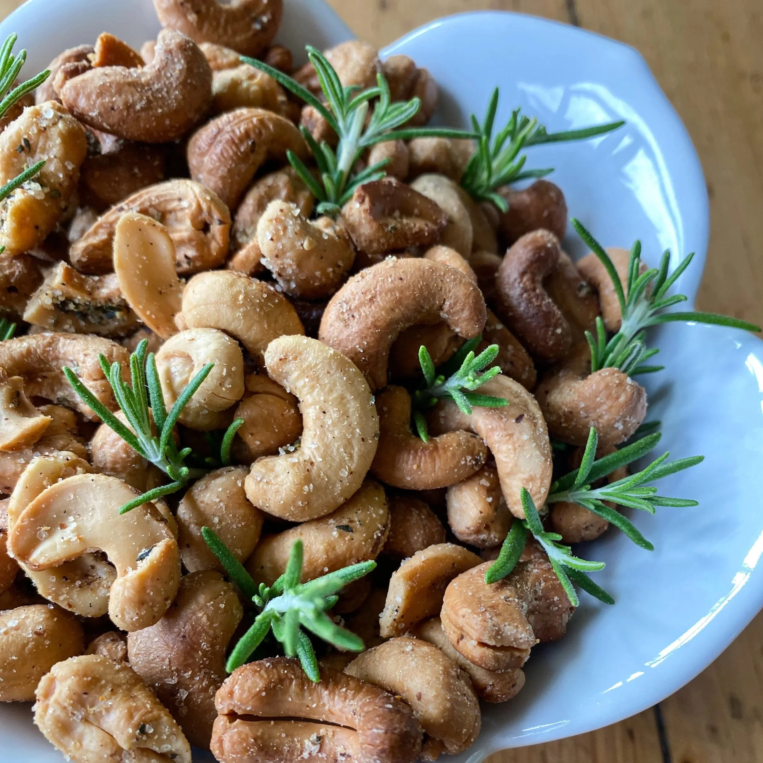 Close-up of a white bowl filled with roasted cashews, garnished with sprigs of fresh rosemary.