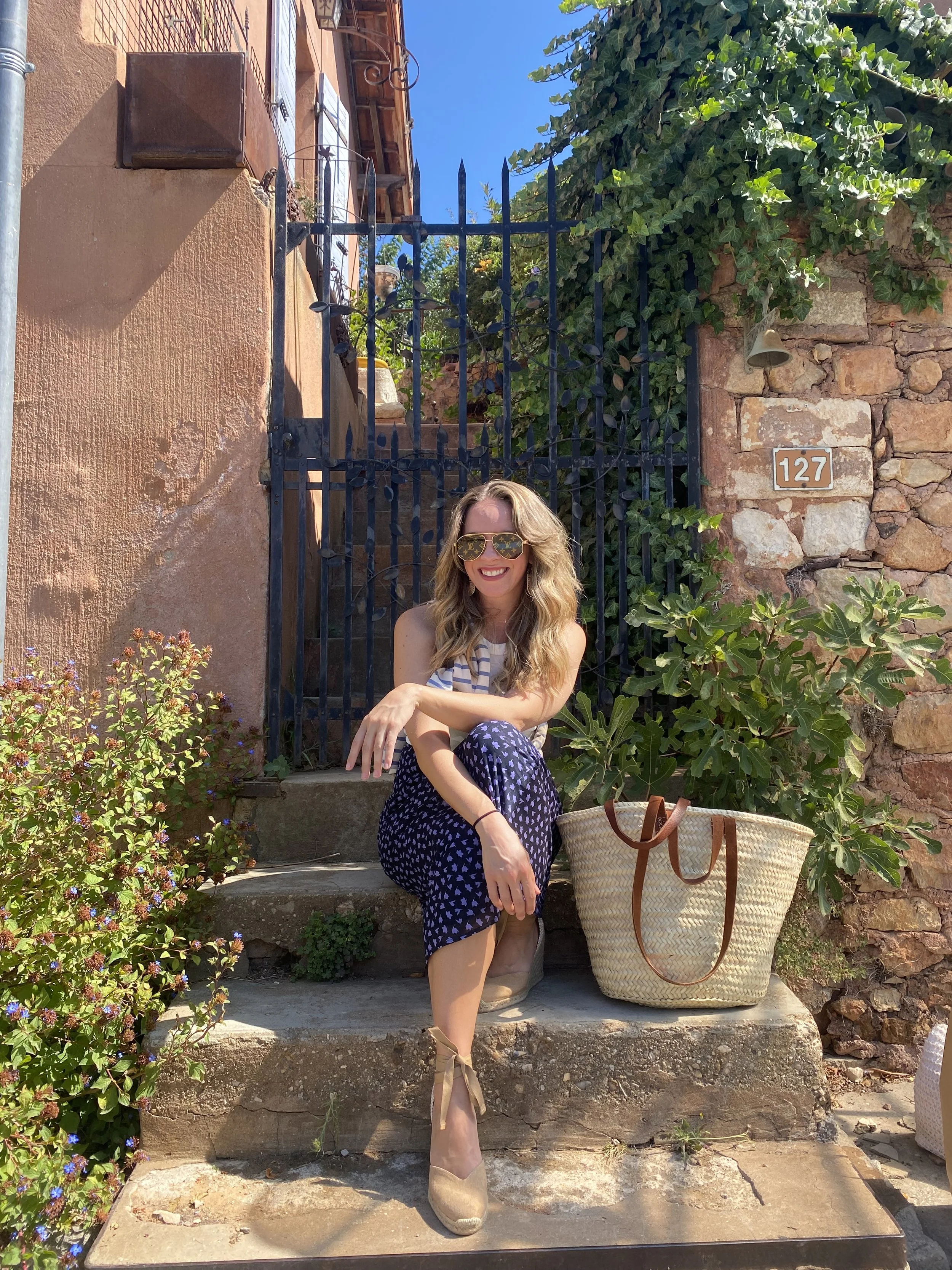 A woman with long blonde hair and sunglasses is sitting on stone steps outside a stone house with a pinkish-orange wall, surrounded by green plants and flowers. She is smiling, wearing a striped top and dark patterned pants, with a large woven handbag nearby.
