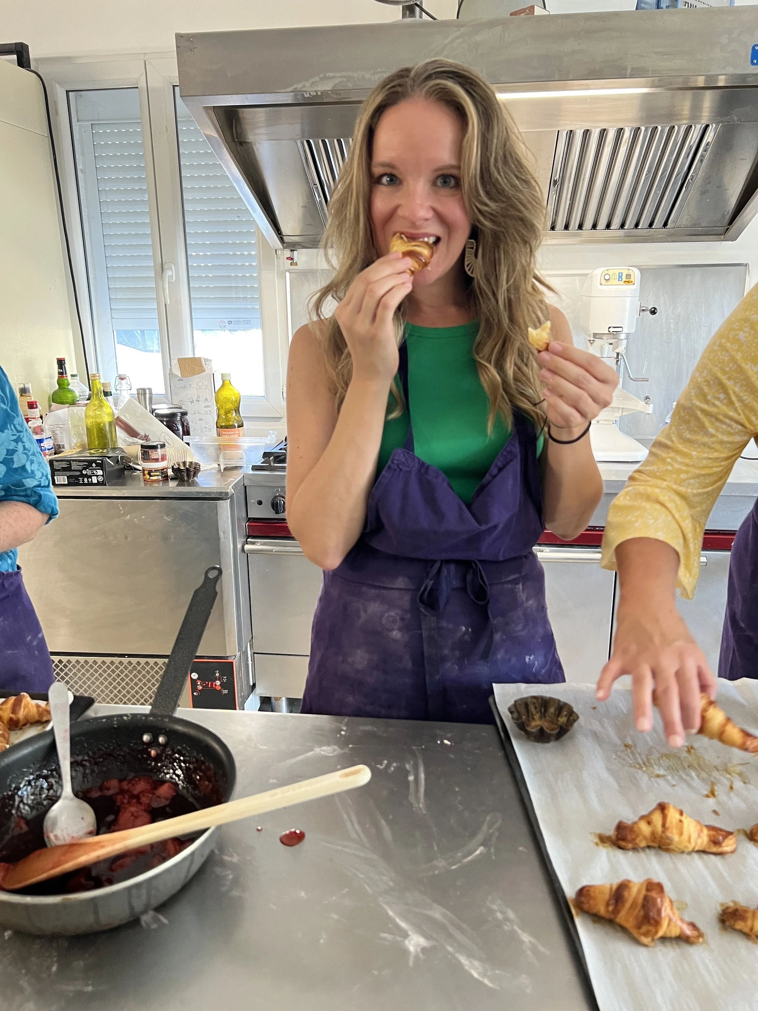 Woman with long wavy hair wearing a green shirt and purple apron tasting food in a commercial kitchen with pastry and berry dish, and cooking utensils.