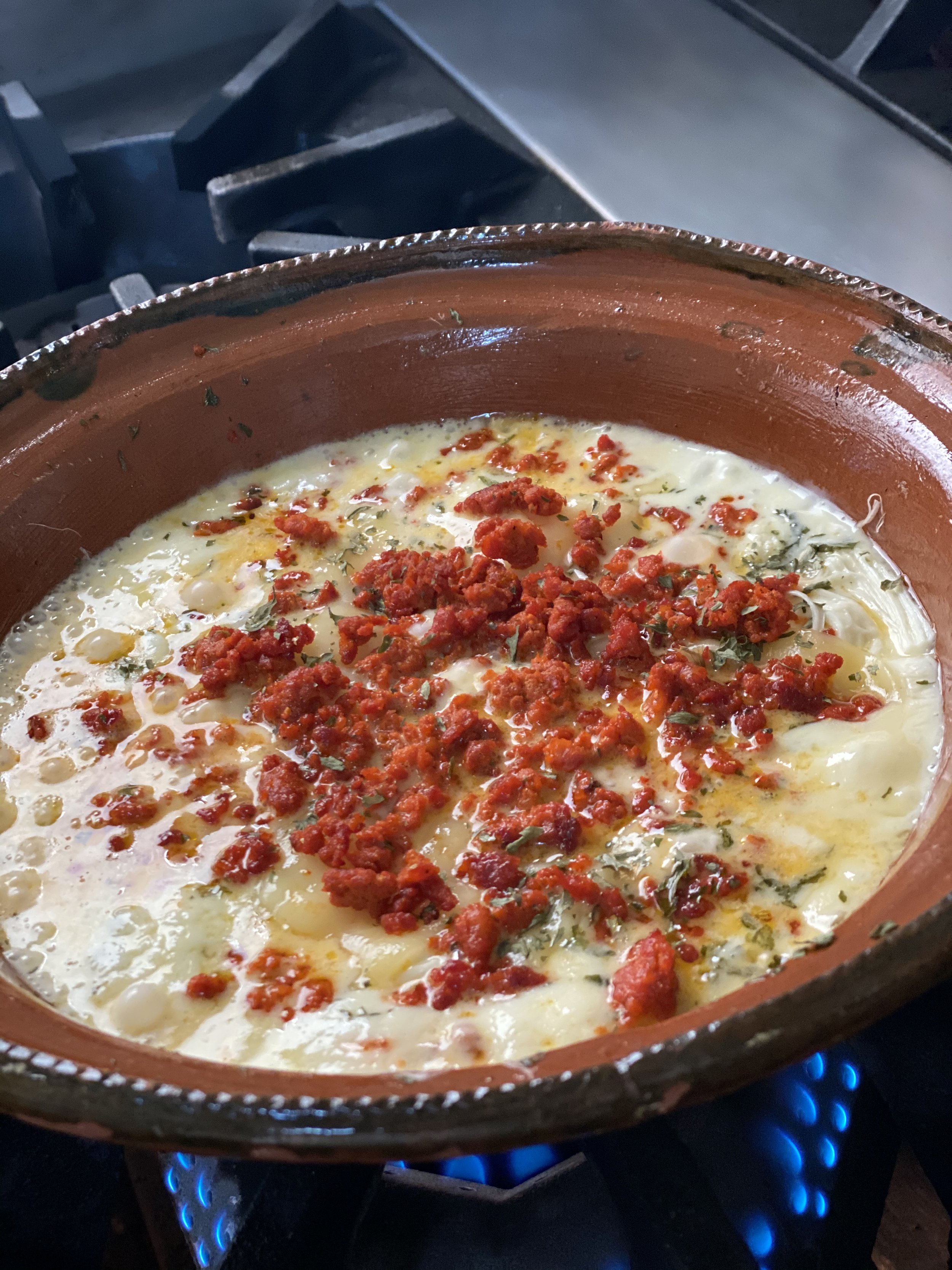 A dish of eggs with melted cheese, topped with crumbled red chorizo sausage and herbs, cooking in a clay pot on a stovetop.