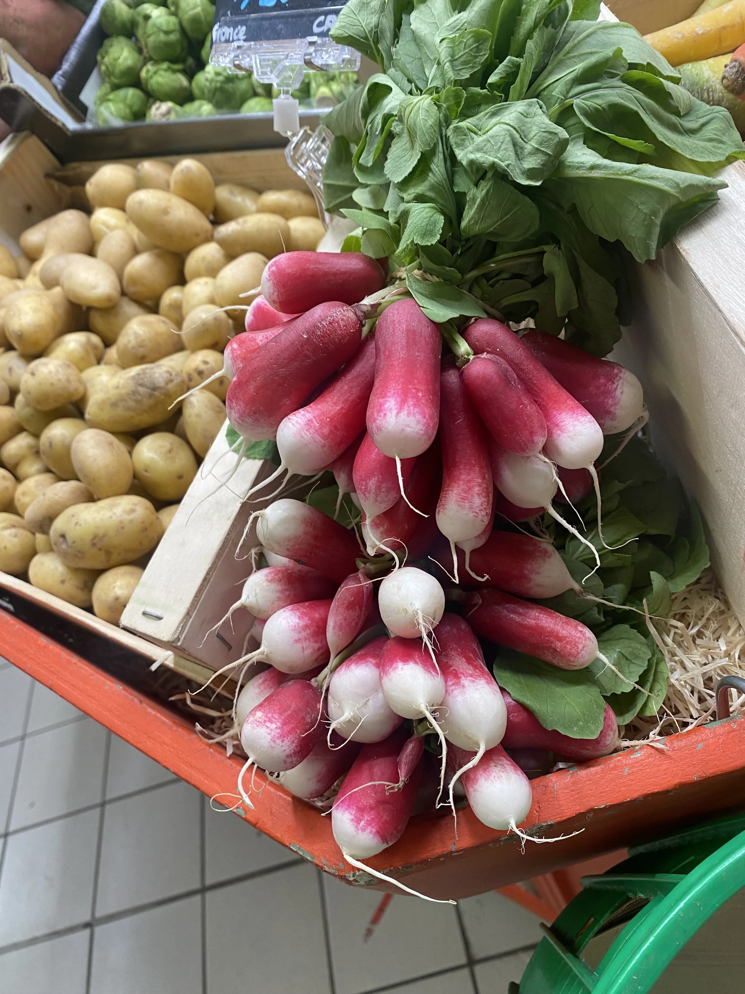 Fresh radishes with green leaves on display at a market, with potatoes and Brussels sprouts in the background.