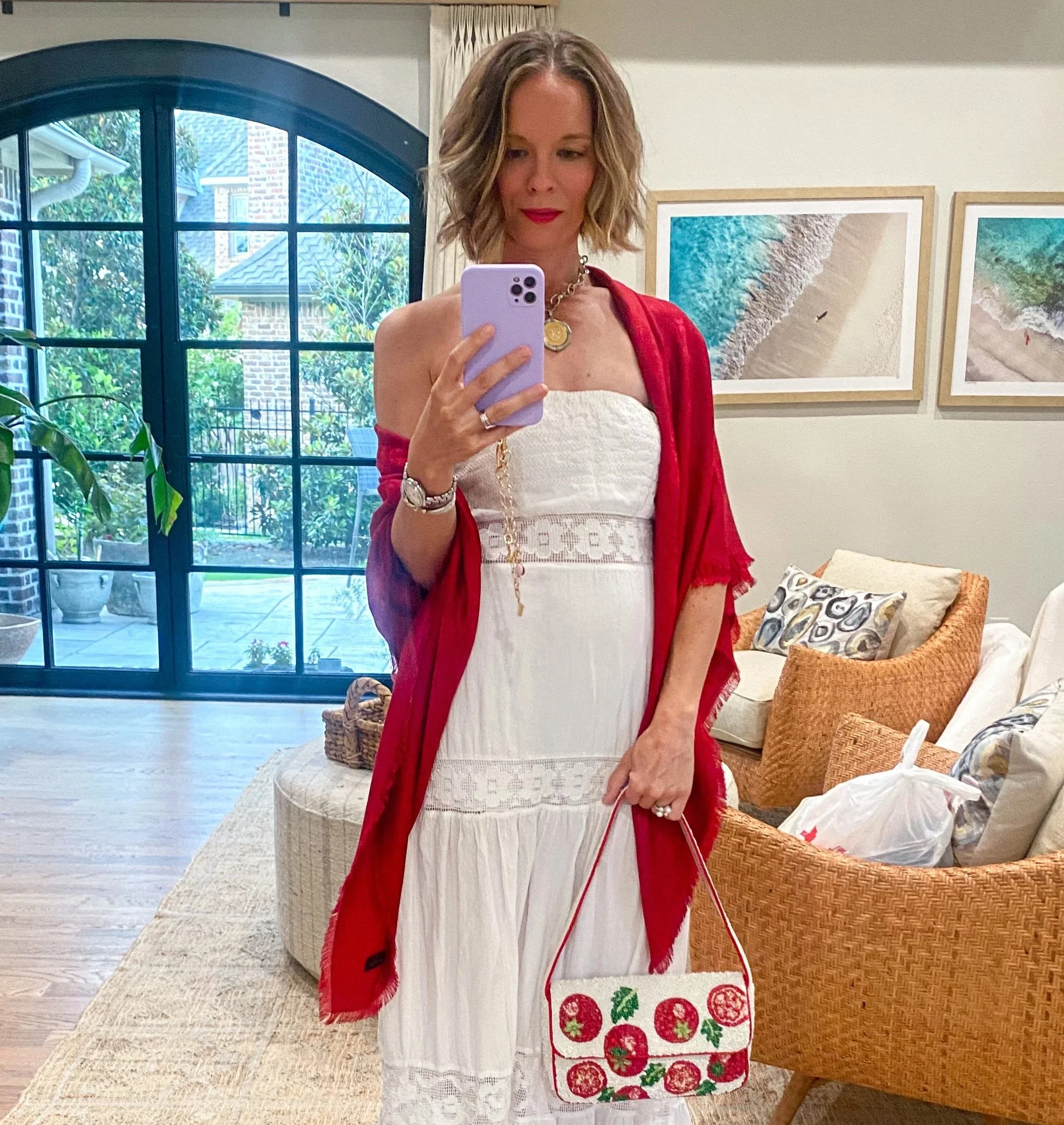 A woman in a white dress and red shawl taking a mirror selfie in a room decorated with beach-themed artwork and furniture. She is wearing jewelry and carrying a tote bag with a strawberry print.
