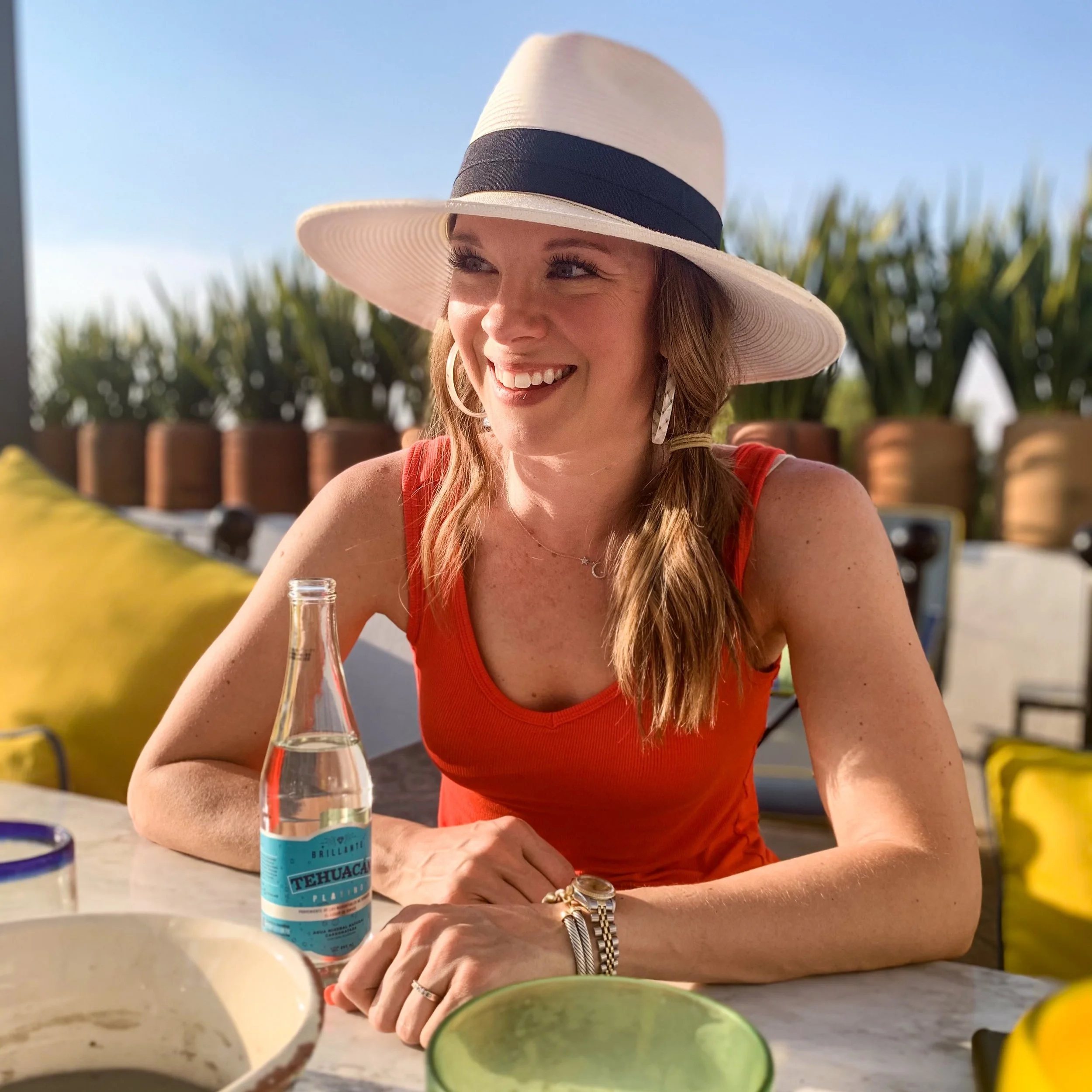 A woman with red hair wearing a large white sun hat with a black band, smiling and sitting outdoors under a clear blue sky. She is near a table with a glass bottle in front of her, surrounded by colorful bowls and cups, with a background of green plants and a wooden fence.