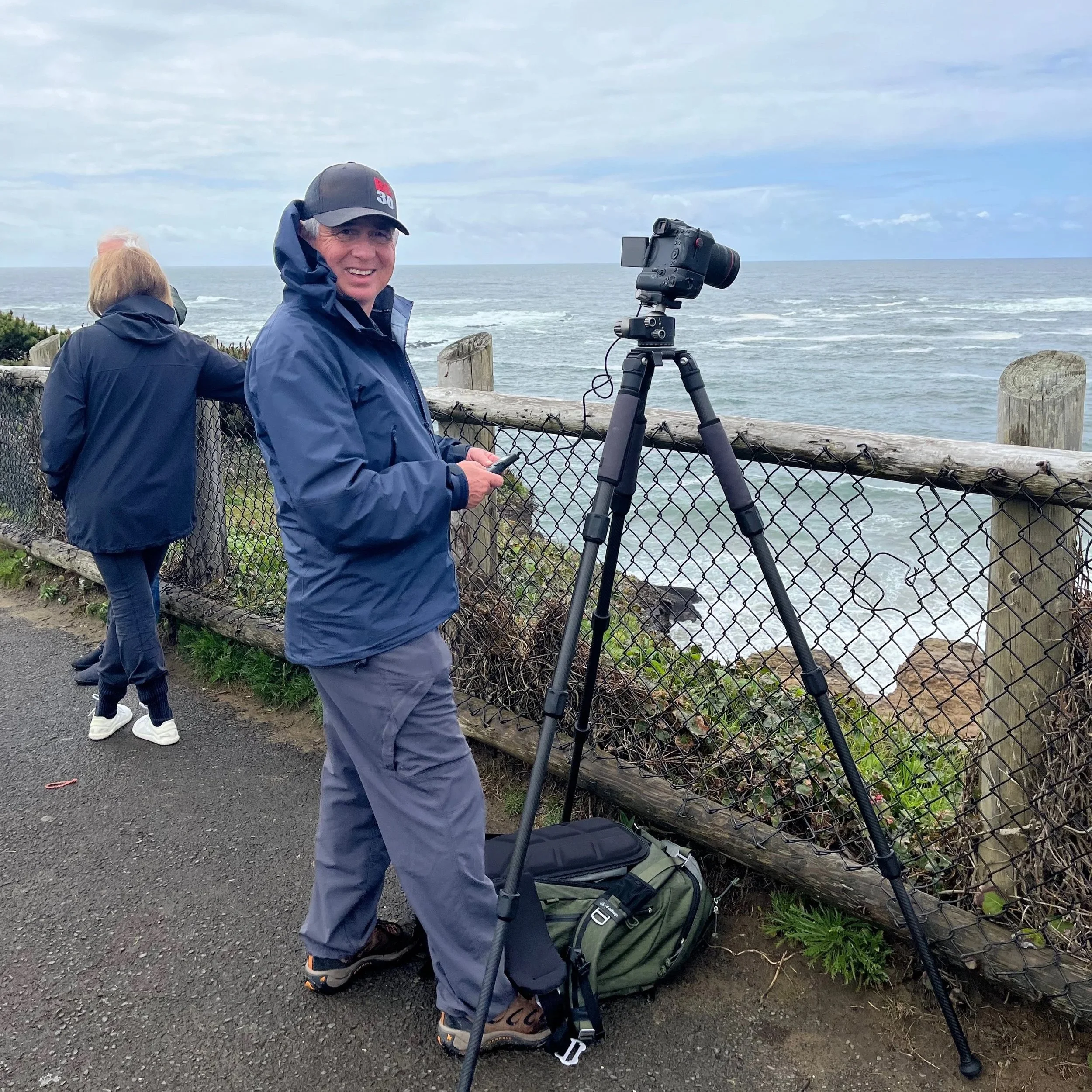 Man in blue jacket and hat standing by a fence at a coastal viewpoint with camera on a tripod, smiling at the camera.