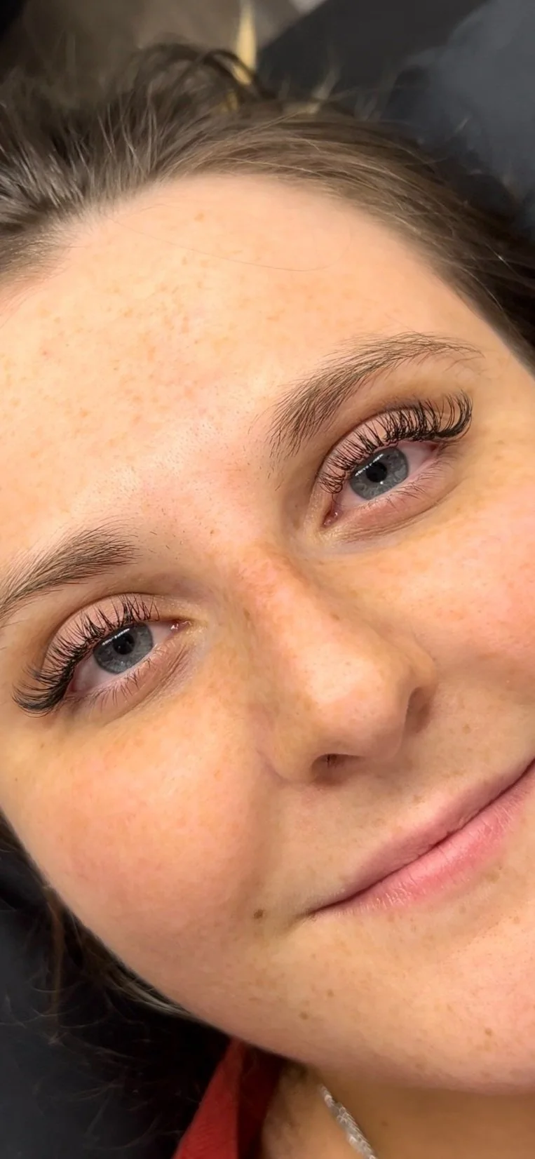 Close-up of a young woman's face with natural freckles, blue eyes, and long eyelashes, lying down and smiling slightly.