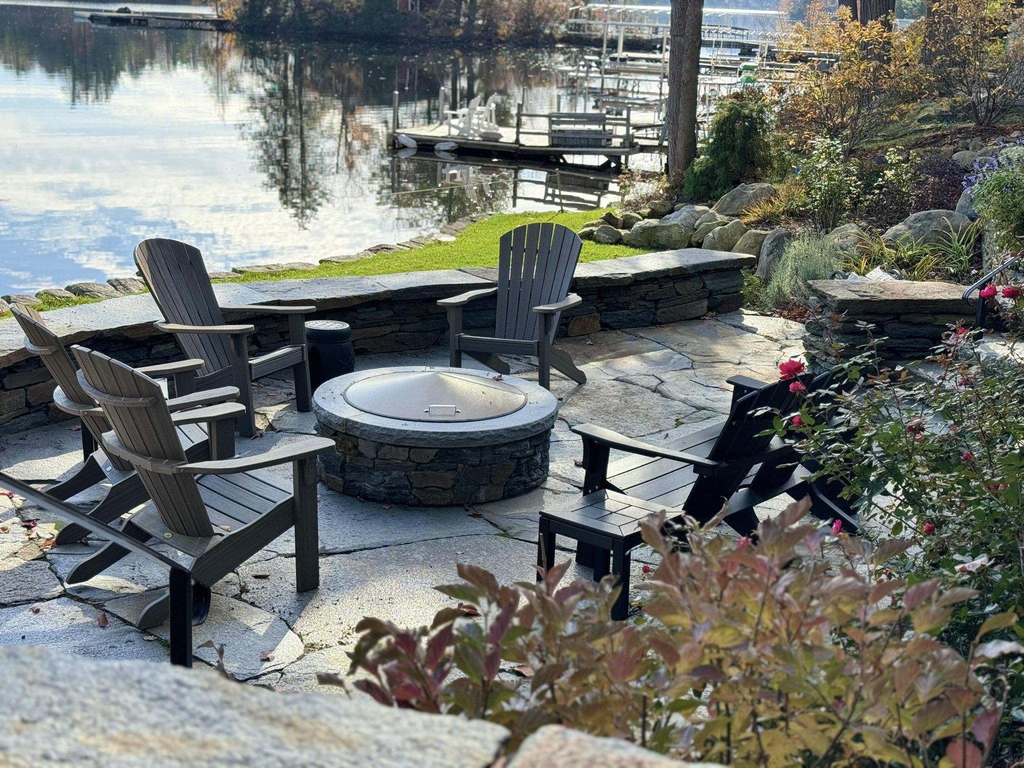 On a calm summer day by the lake, laughter and conversation fill the air as friends gather on a Goshen stone patio. Seated comfortably around a fire pit, the warmth of flames dances in sync with the serene waters