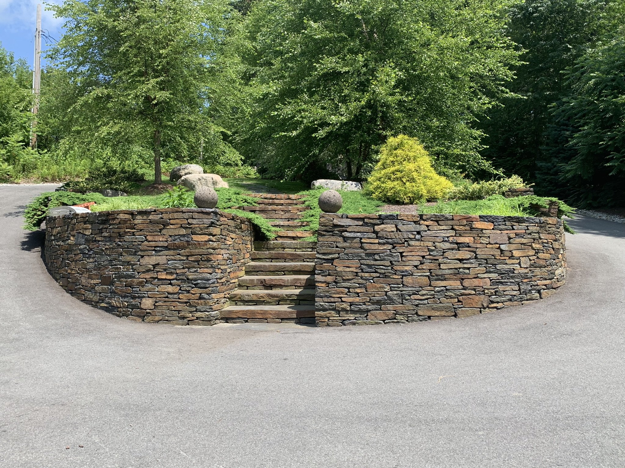 A skillfully crafted Goshen stone retaining wall graces the driveway as a centerpiece, accented with flourishing trees and shrubs
