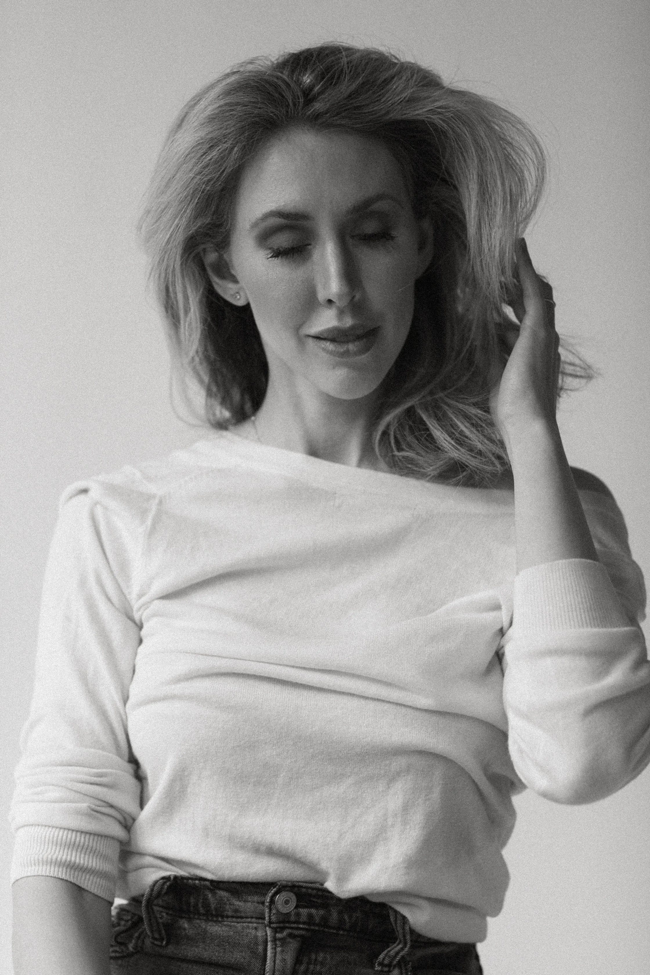 A woman with long hair and closed eyes, wearing a white long-sleeved shirt and jeans, poses against a plain background with her hand touching her hair.
