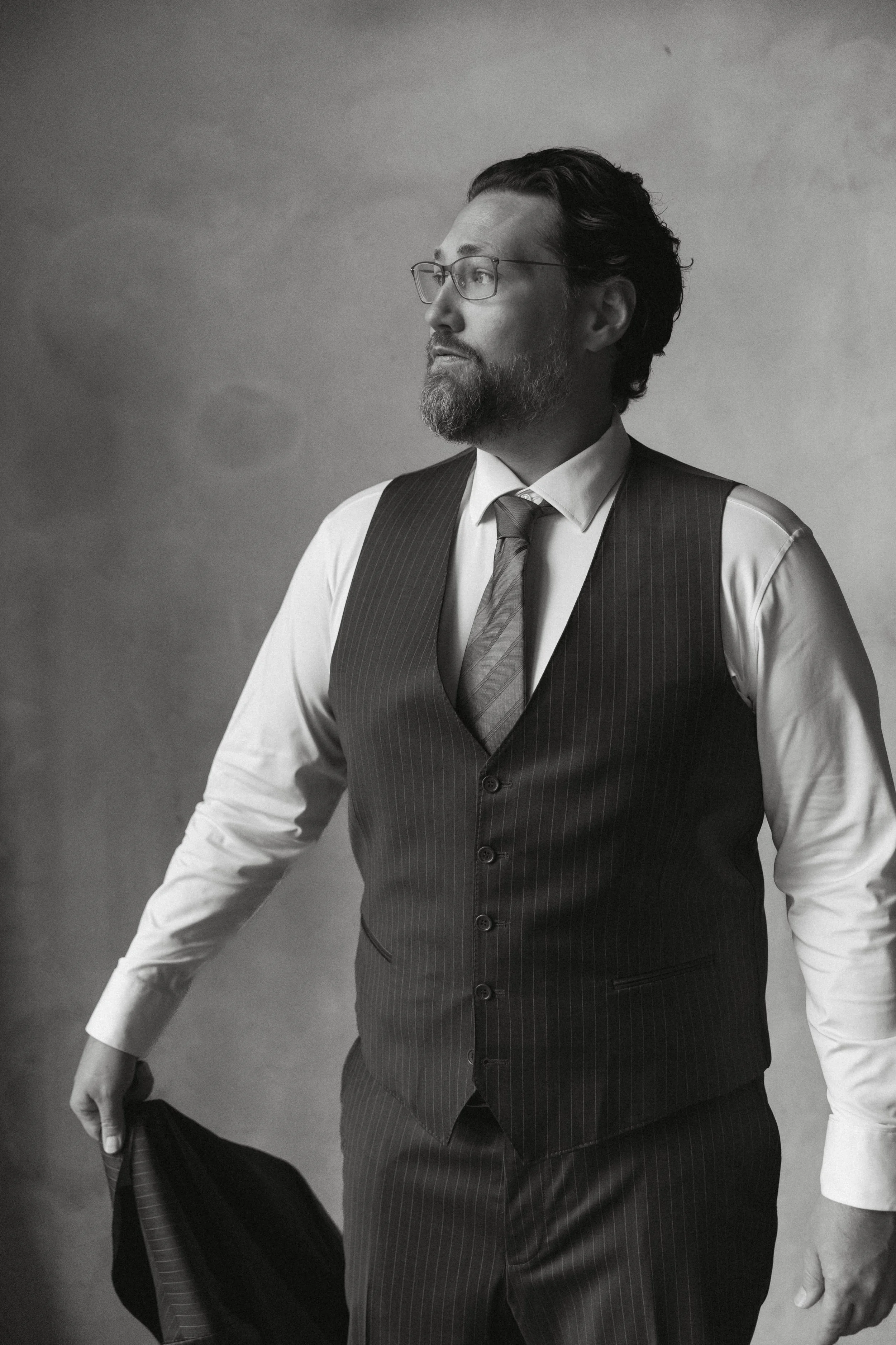 Black and white headshot of man in vest and tie holding jacket with editorial studio lighting
