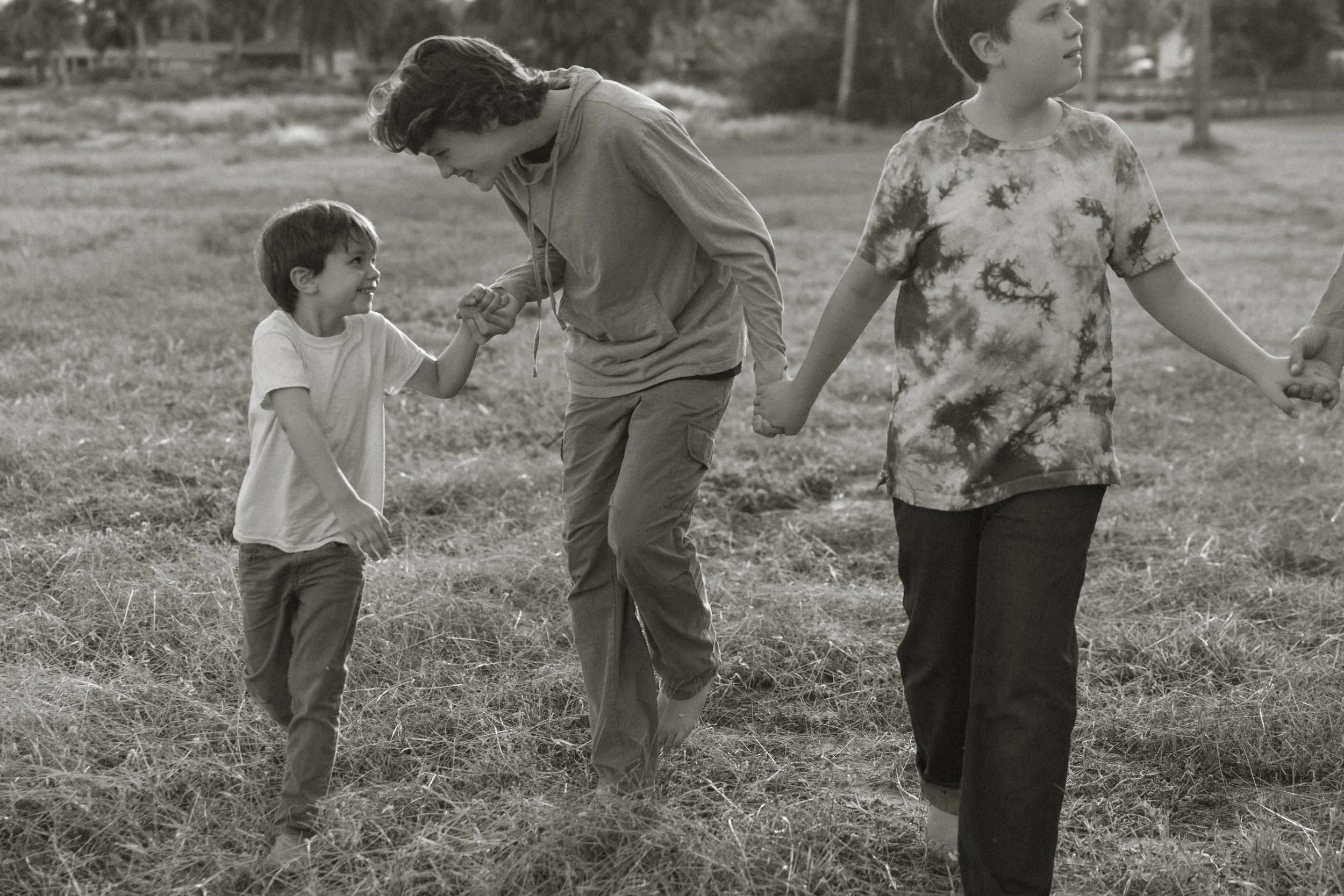 Older brother holding hands with his younger brother while walking and smiling together outdoors in a grassy field, candid family photography session in Orlando.