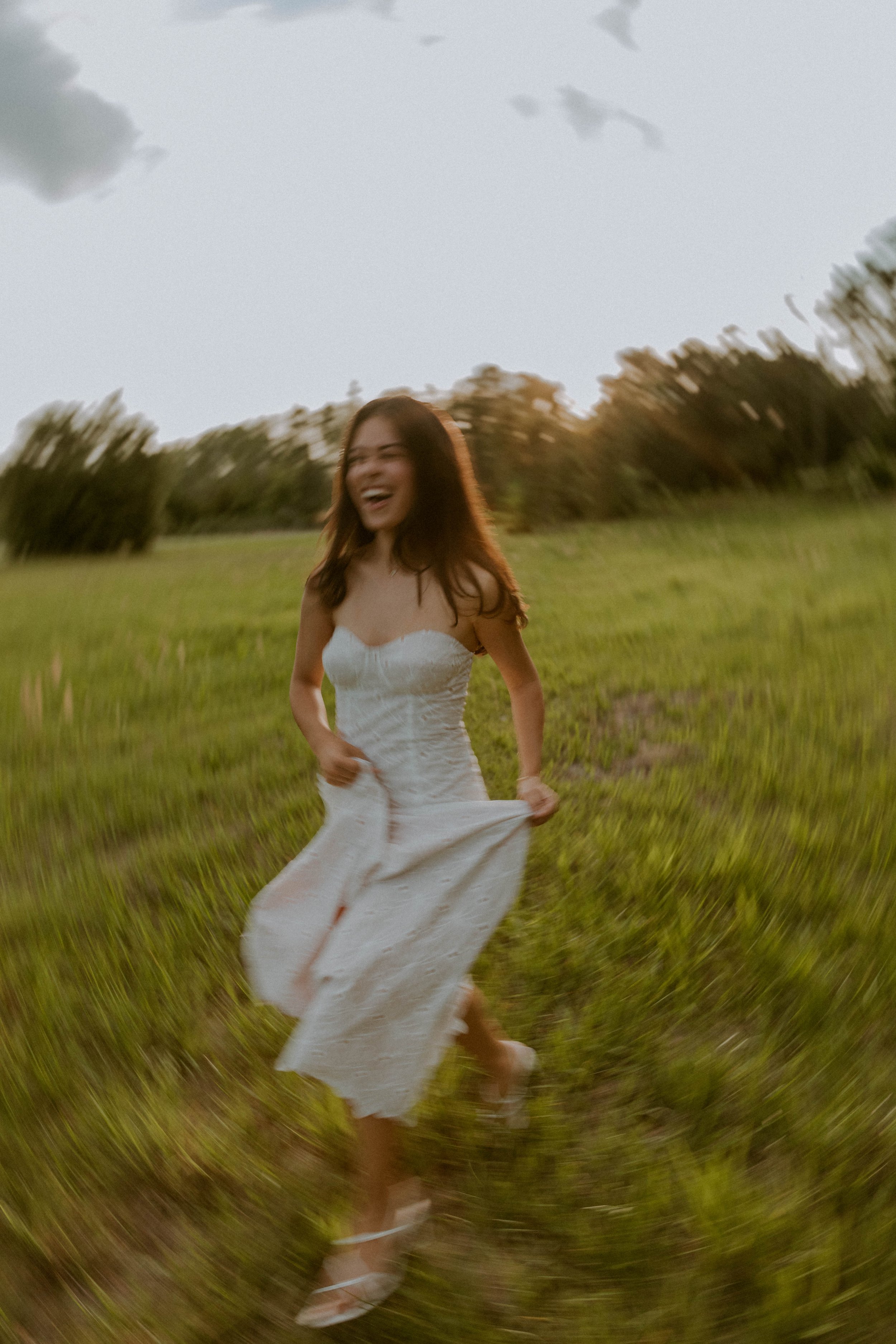 A woman in a white dress running through a grassy field with trees in the background during sunset, with a motion blur effect.