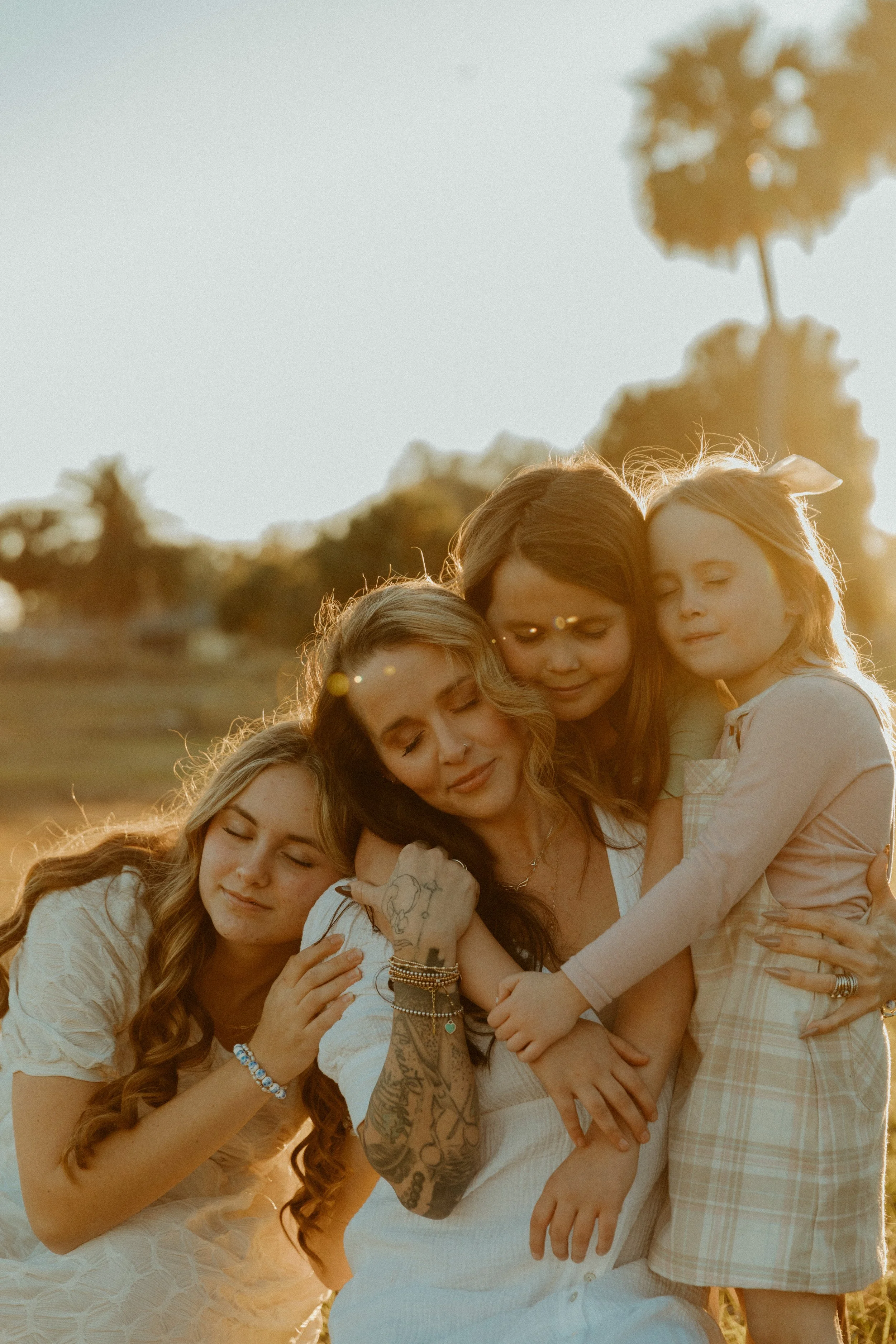 Mother embracing her three daughters in warm golden light, eyes closed and leaning into each other during a relaxed outdoor family photography session.