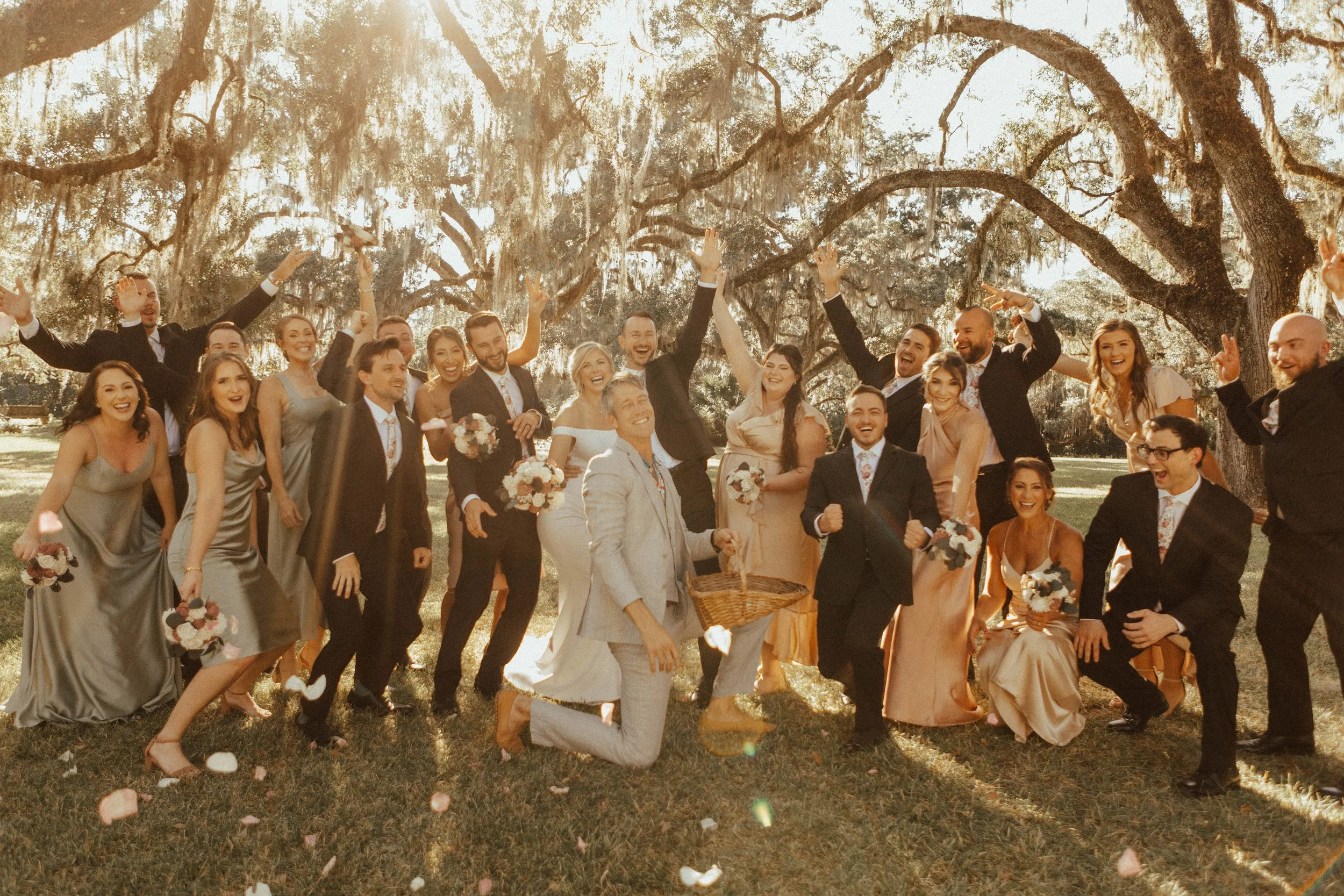 A large group of smiling people, dressed in formal attire, celebrating outdoors under large, sprawling tree branches. Some hold flower bouquets, and the scene is illuminated by sunlight, creating a joyful and festive atmosphere.