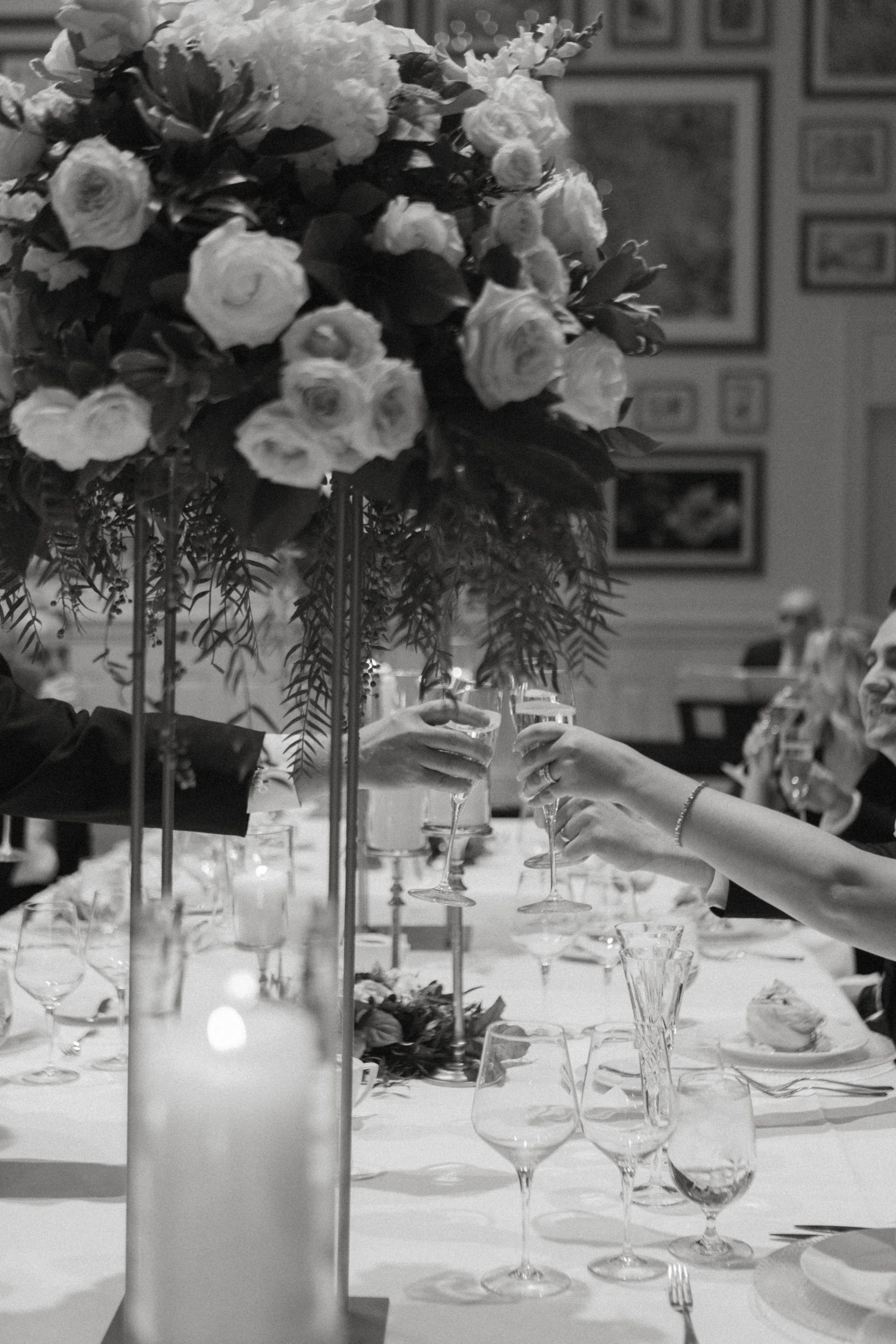 A formal dinner table with a large floral centerpiece, wine glasses, and place settings. People are toasting with champagne glasses amidst a background of framed pictures on the wall.