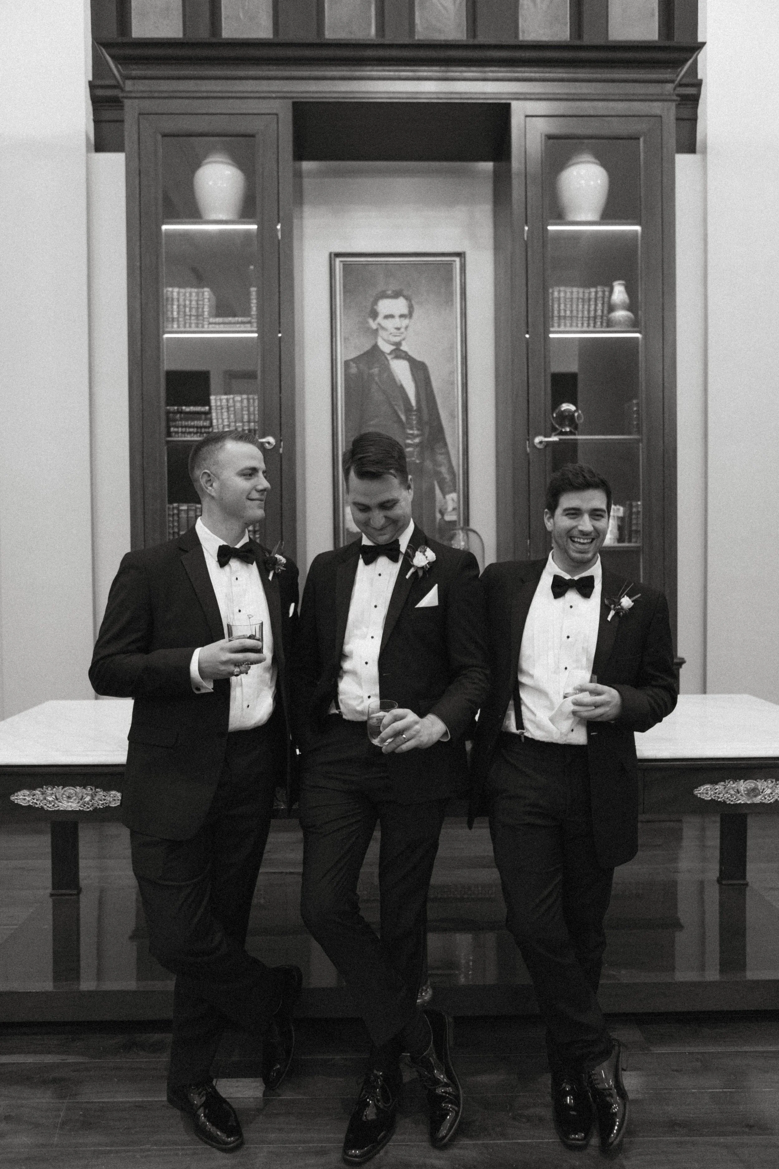 Three men in tuxedos with bow ties smiling and holding glasses, standing in front of a framed photo of a man in formal attire, with a scholarly setting in the background.