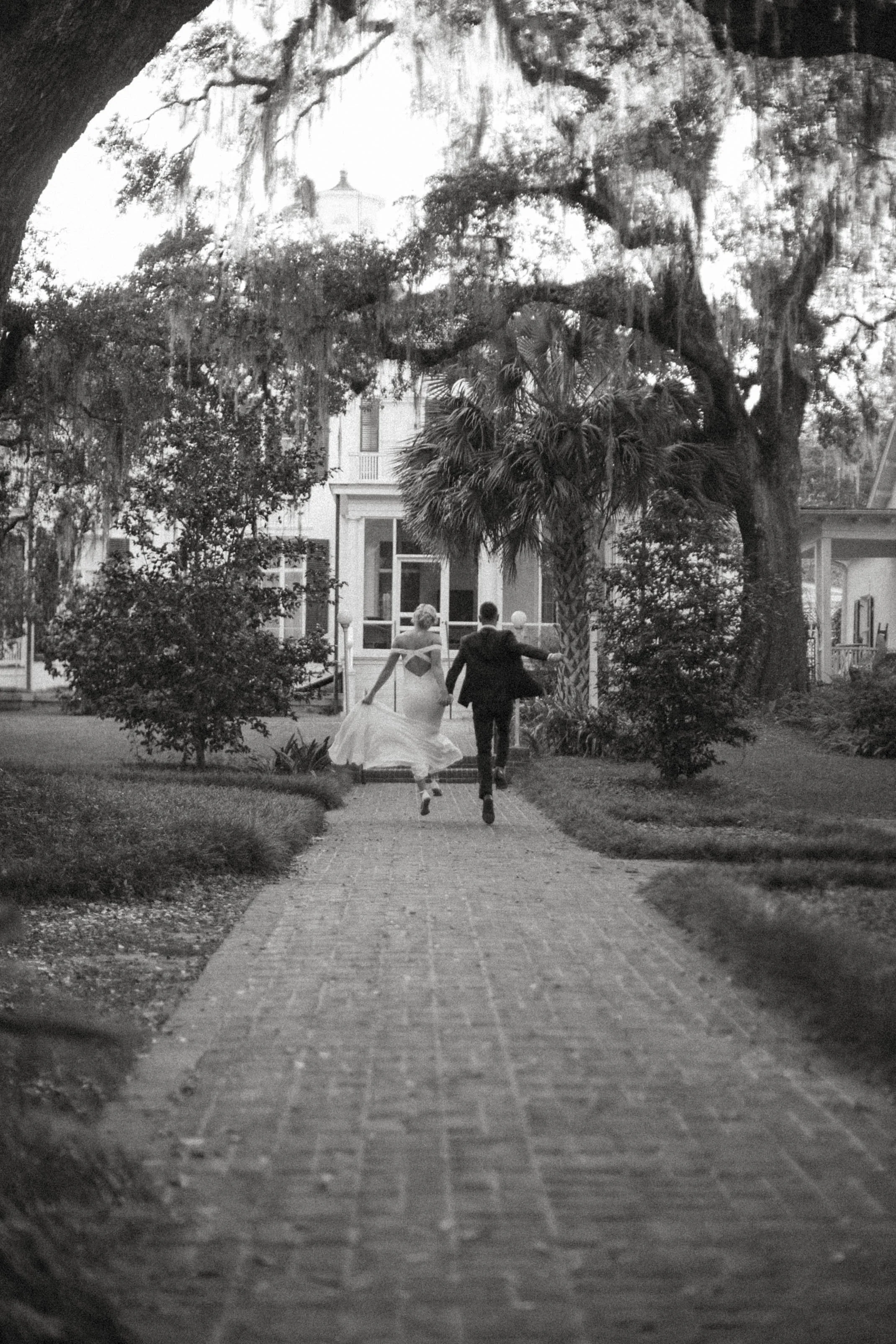 Black and white photo of a newly married couple walking away from the camera on a paved pathway surrounded by trees and bushes, with a large tree overhead and a building in the background.