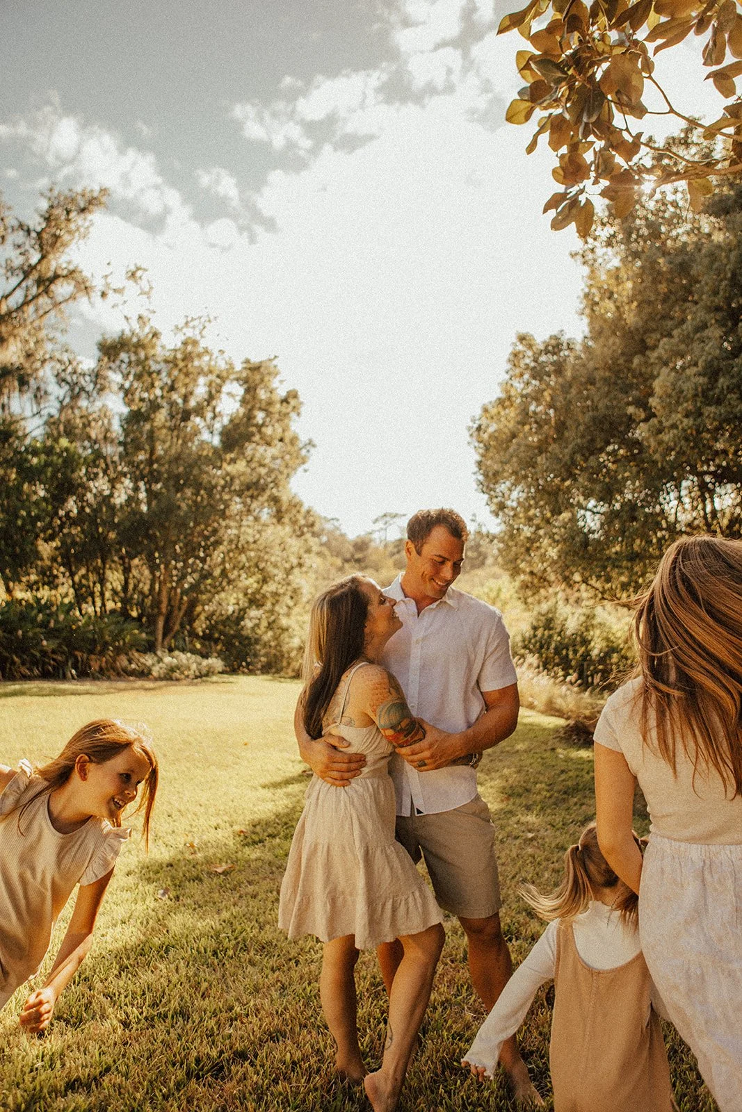 A group of people, including two women and two girls, enjoying a sunny outdoor gathering in a park with trees and grass. A man and a woman are central, embracing and smiling at each other. The women and girls are wearing light-colored dresses, and th