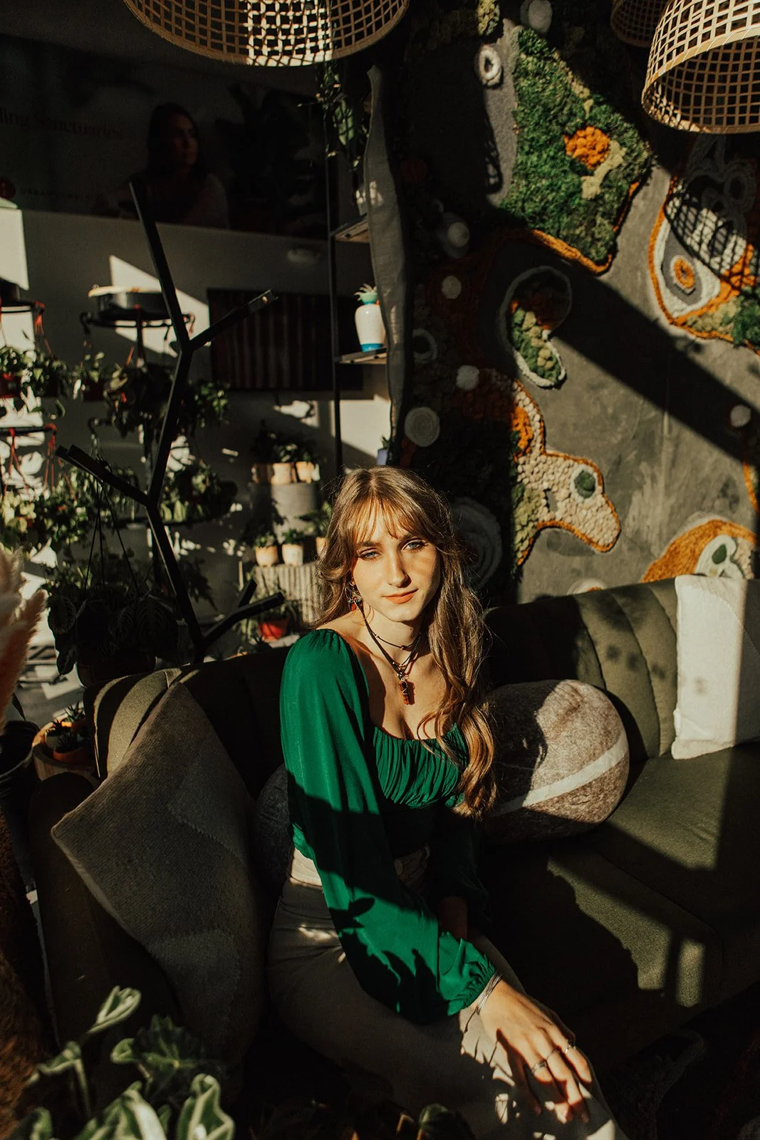 A young woman with long wavy hair sitting on a dark green sofa in a room filled with plants and artwork. She wears a green dress and necklaces, with sunlight casting shadows on her face and body.