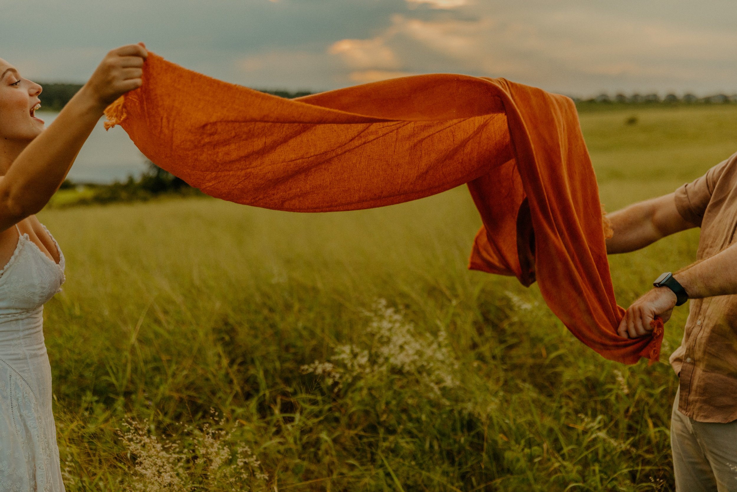 A woman on the left smiling and holding an orange fabric in a field, with another person on the right holding the other end of the fabric; the sky is cloudy with a hint of sunset.