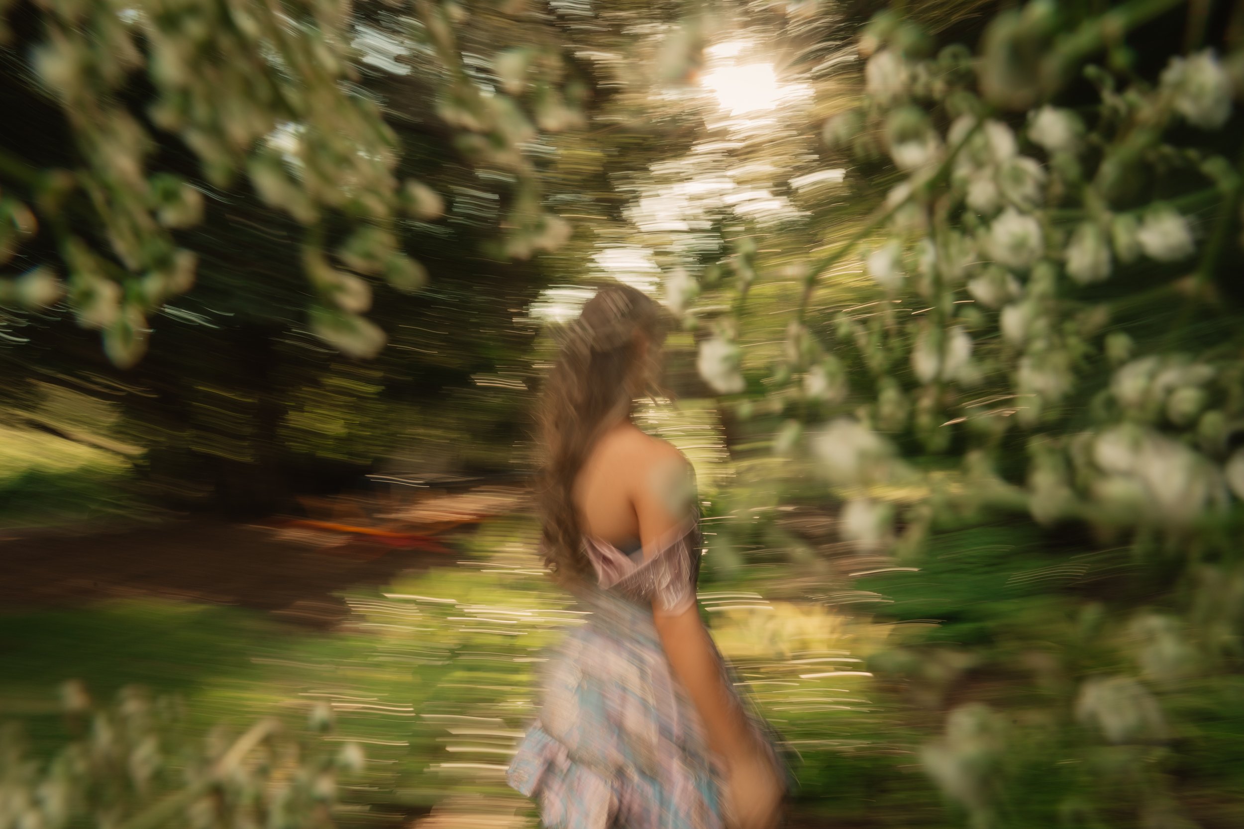 A person with long hair and a dress jogging or walking outdoors among a lush green garden or forest with flowering plants, with sunlight filtering through the trees.