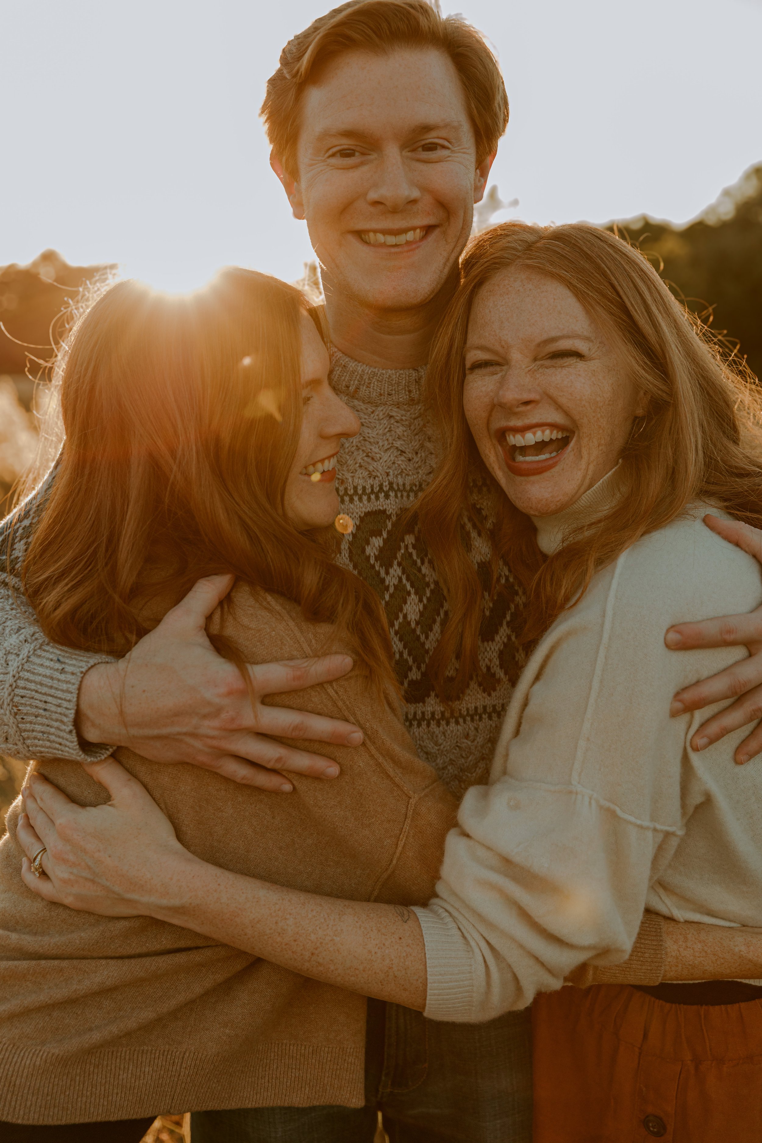 A group of three friends hugging each other during sunset, smiling and laughing, outdoors with trees and sky in the background.