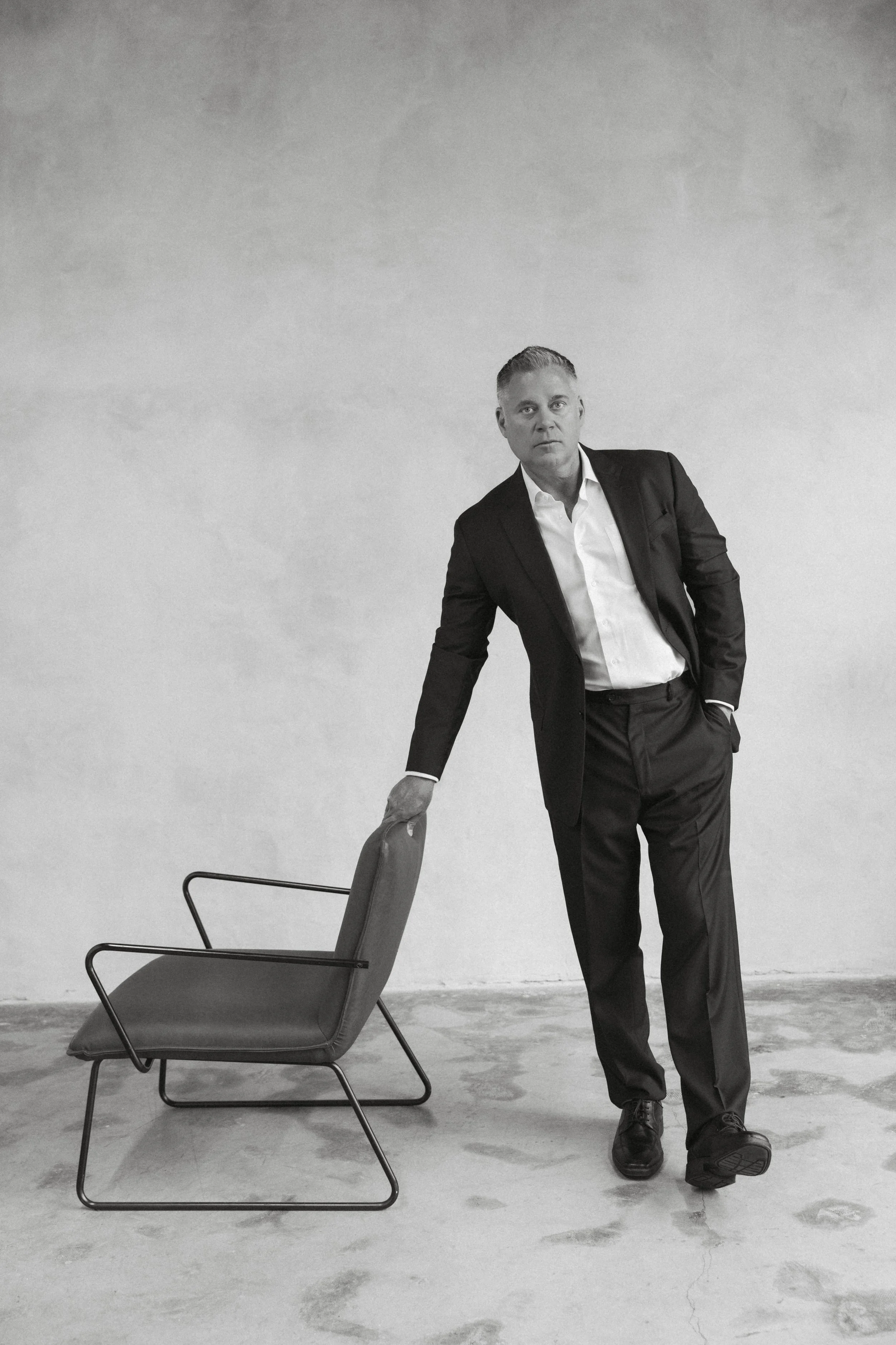 Black and white branding portrait of man in suit leaning on chair in minimalist studio setting