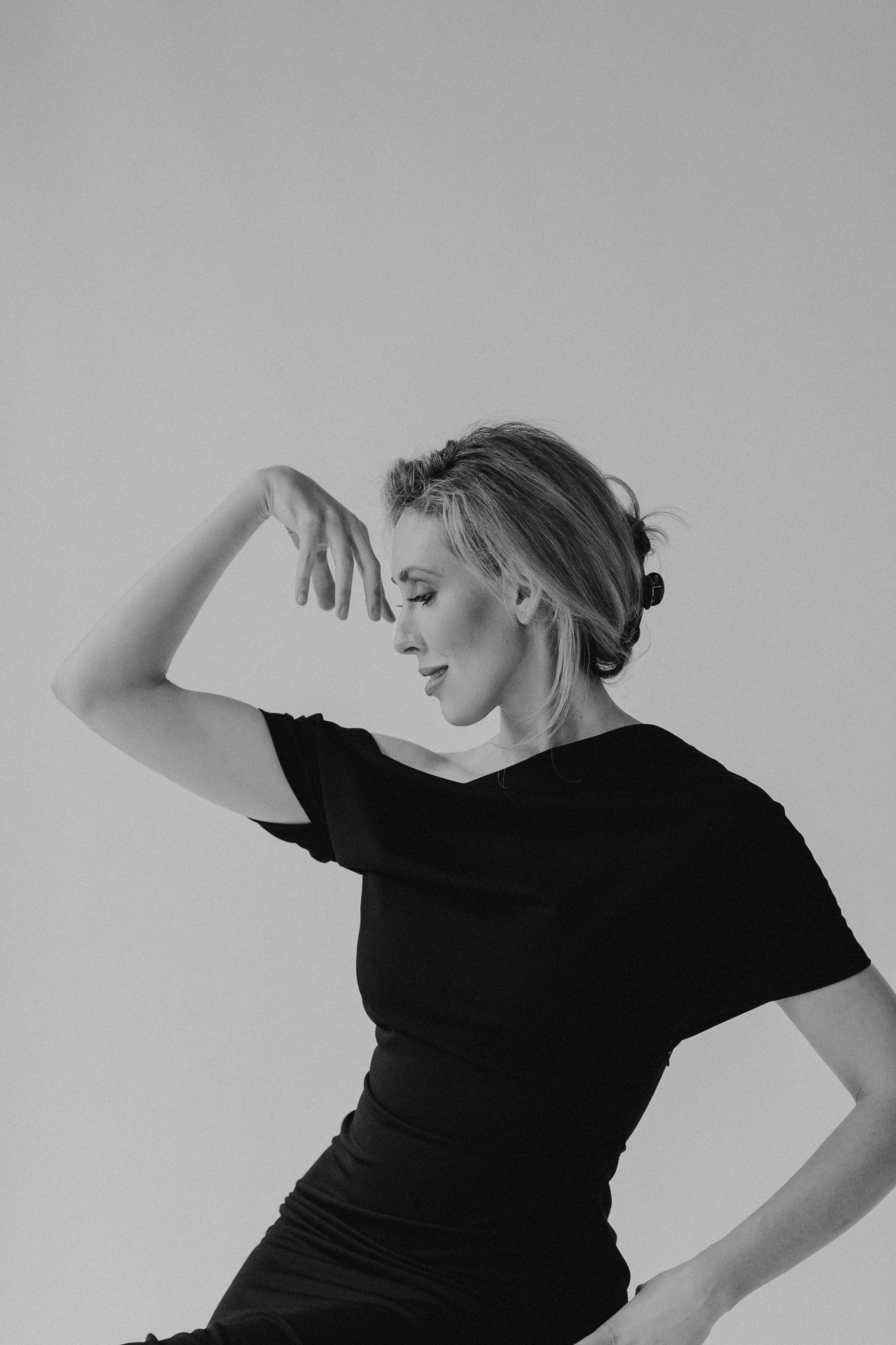 A woman with light-colored hair styled in a side part, wearing a black top, is posing with her eyes closed and arm raised, touching her forehead, against a plain light background.