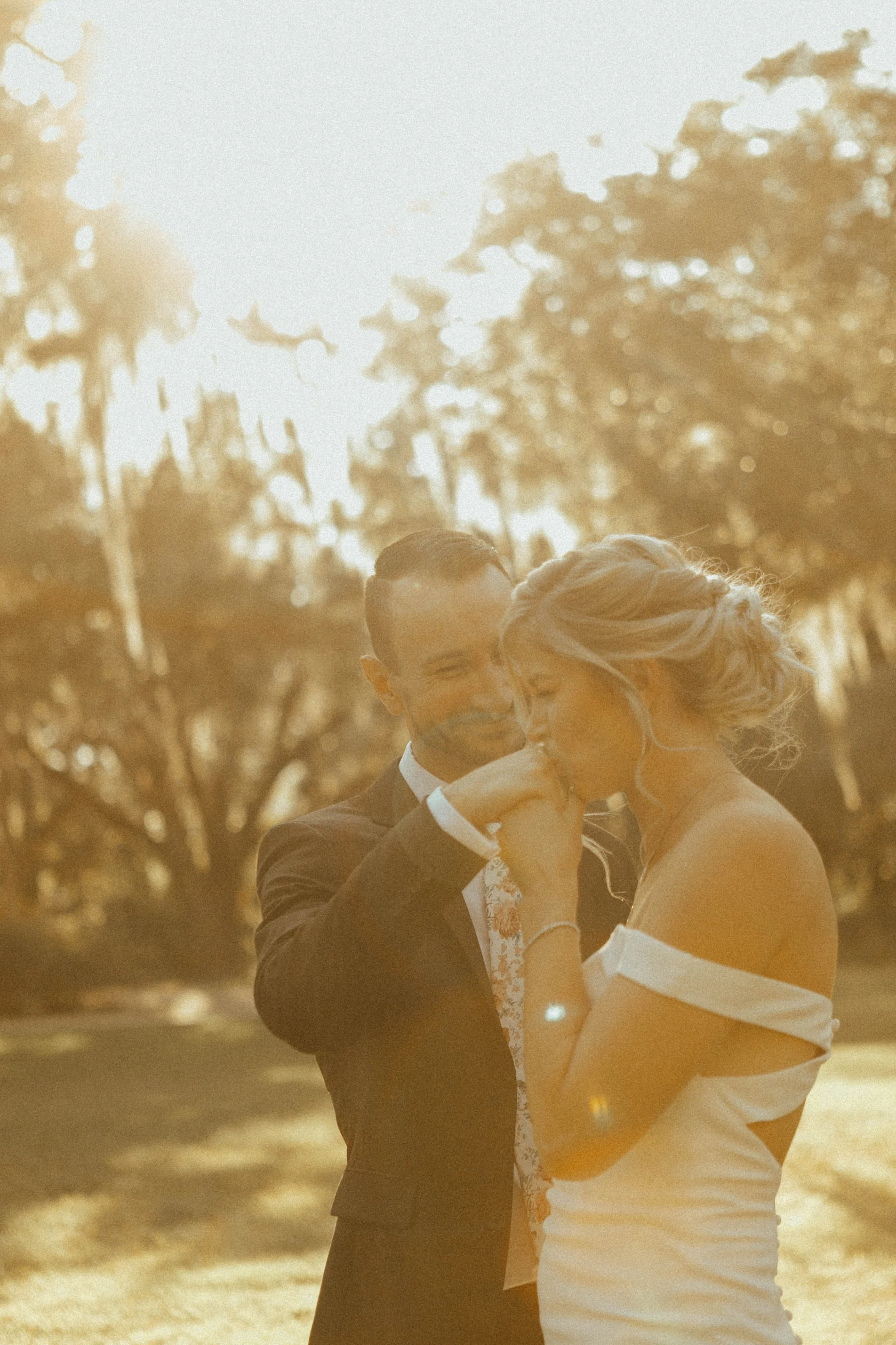 A couple dressed in formal attire sharing an intimate moment outdoors during sunset, with sunlight filtering through trees in the background.