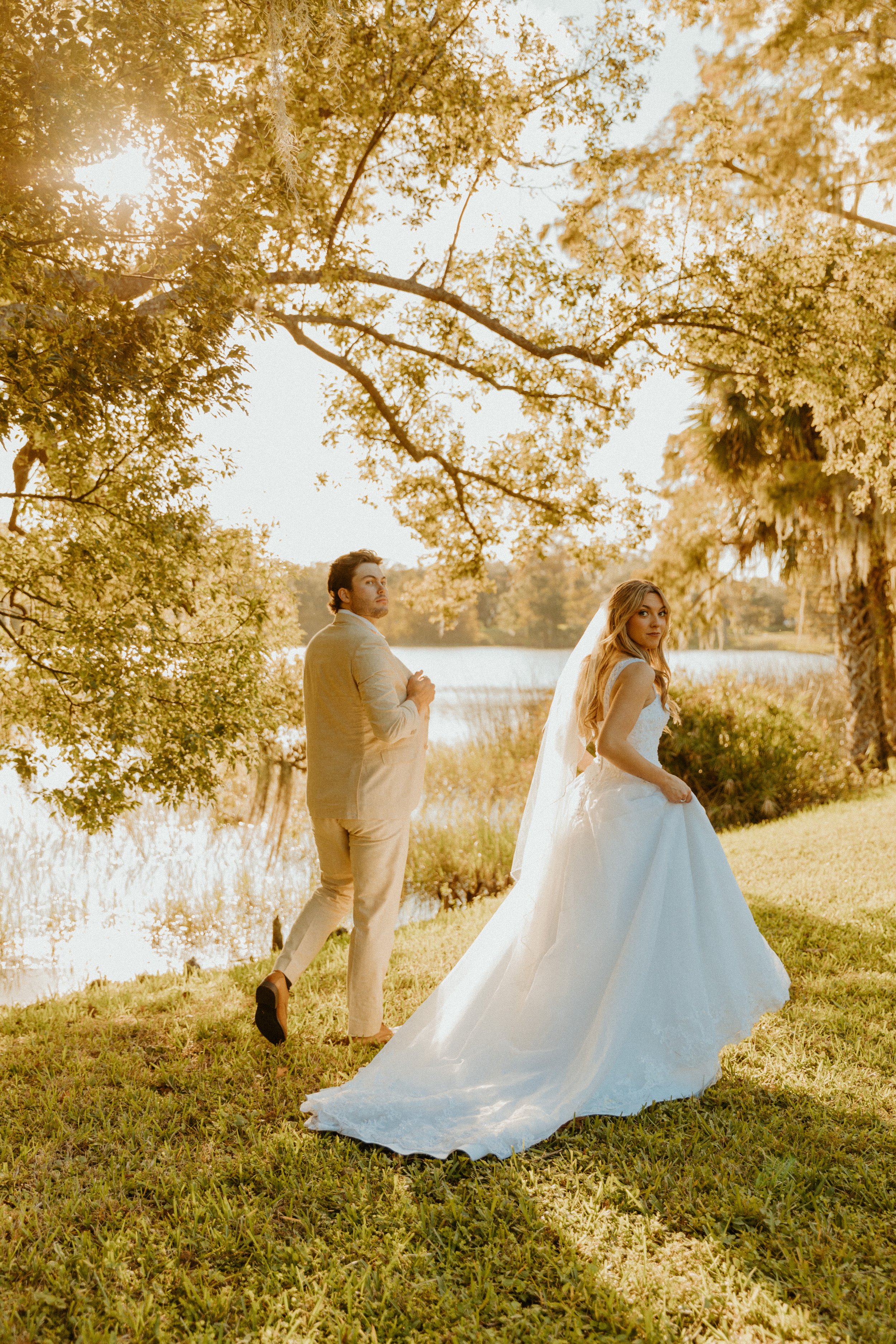 A bride and groom outdoors near a lake with trees. The groom, in a beige suit, is turning away, and the bride, in a white wedding dress, is looking at the camera. The scene is lit with warm, golden sunlight.
