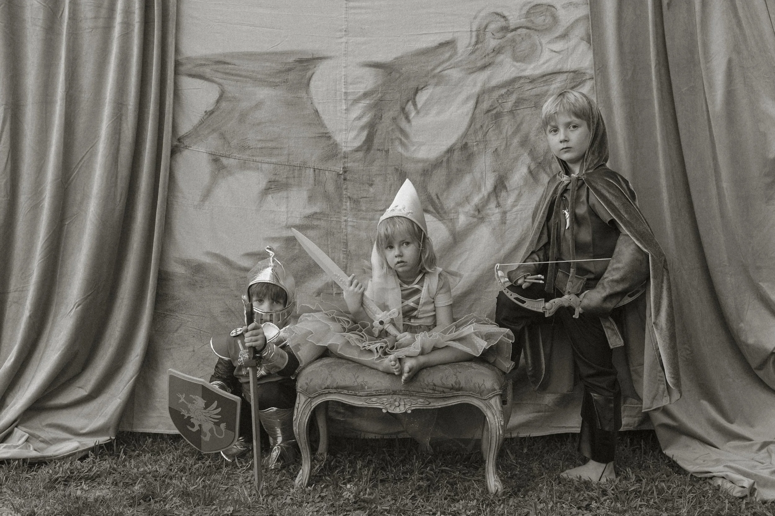 Children dressed as knights and princesses in a castle-themed setting with a backdrop of a mountain and a dragon, with curtains framing the scene.