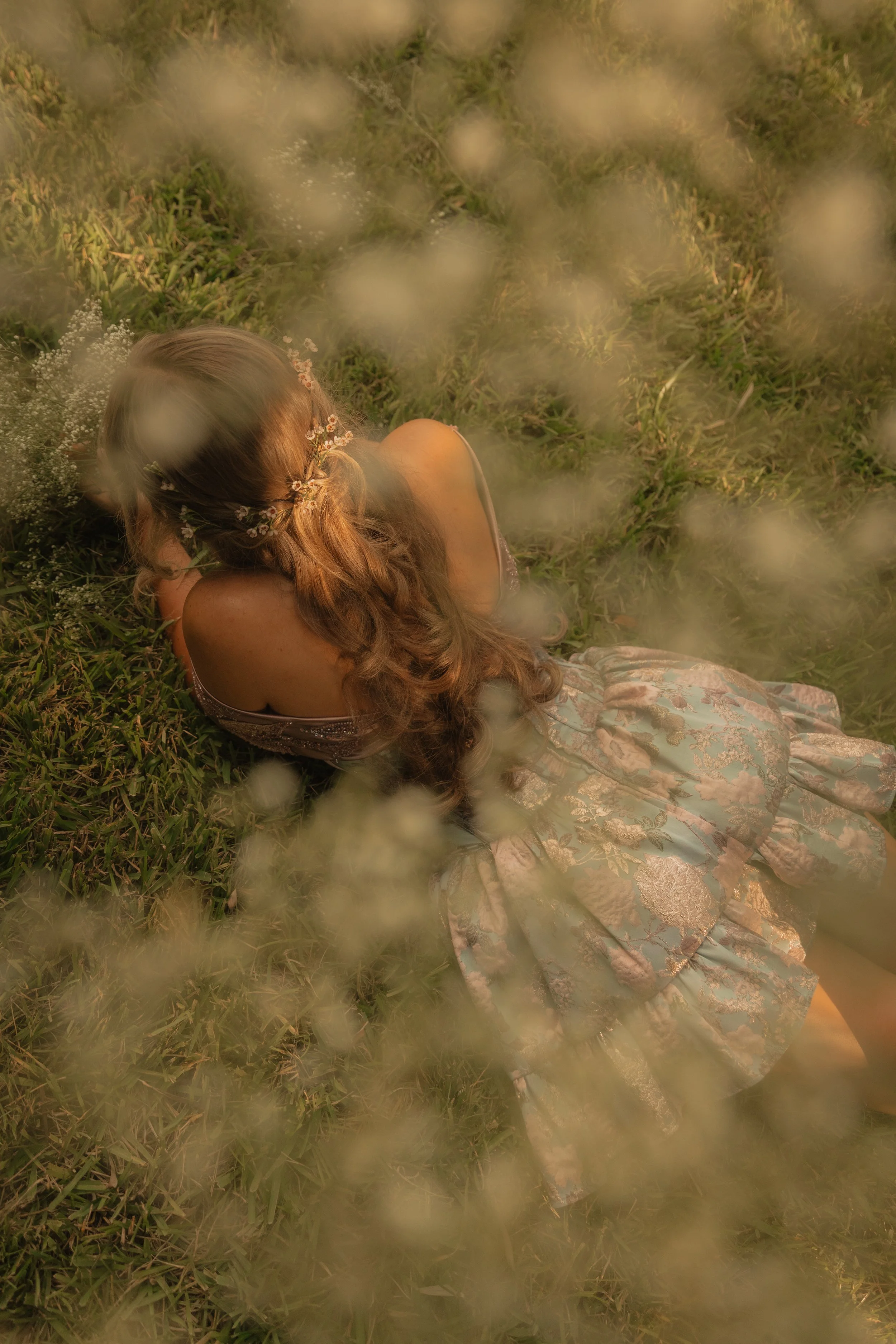 A woman with long, wavy hair lying on grass in a field, surrounded by blurred white flowers and greenery, wearing a floral dress and a floral headband.