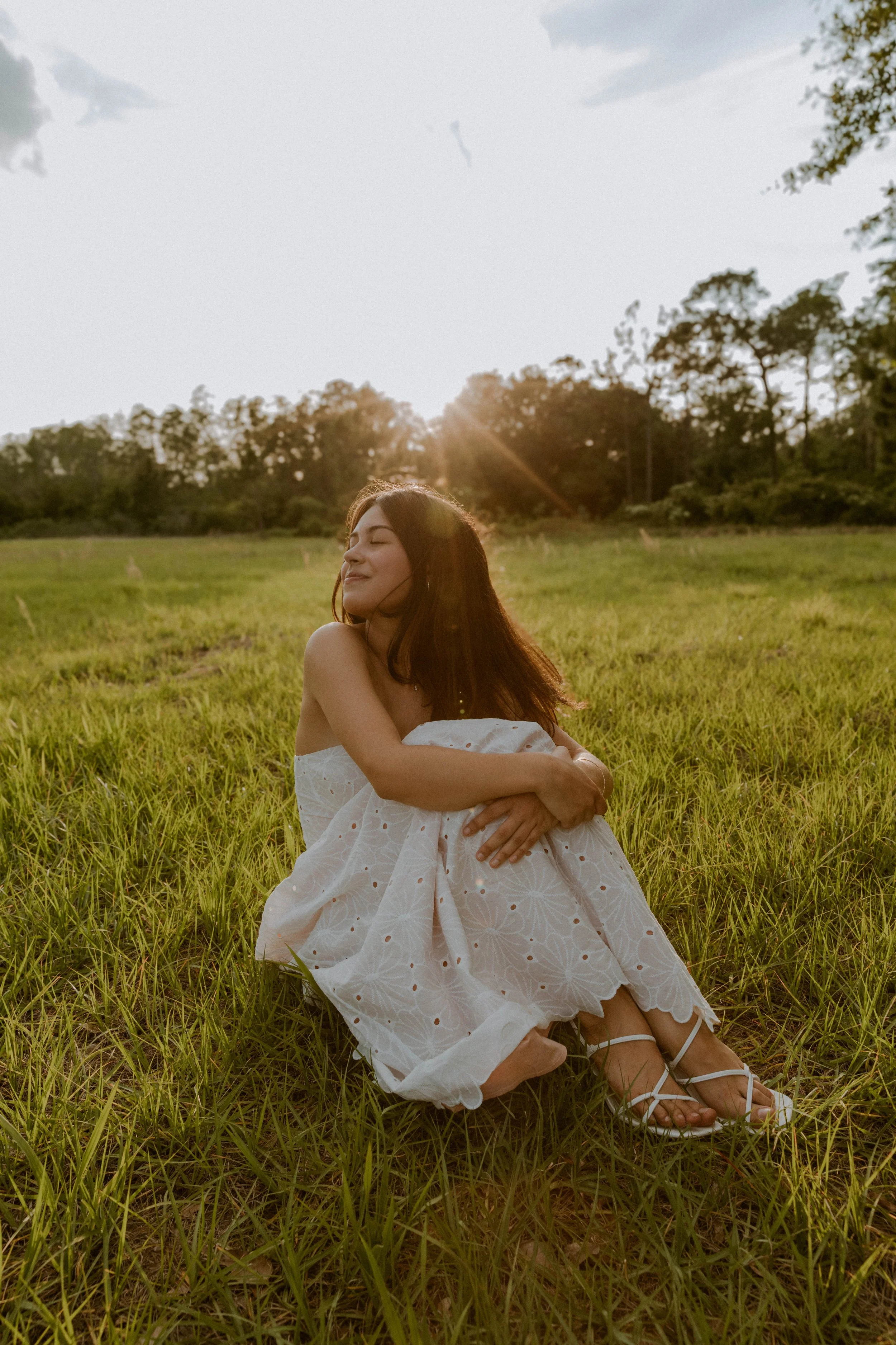 A woman sitting on a grassy field at sunset, wearing a white dress with eyelet details and white sandals, with her eyes closed and a peaceful expression.