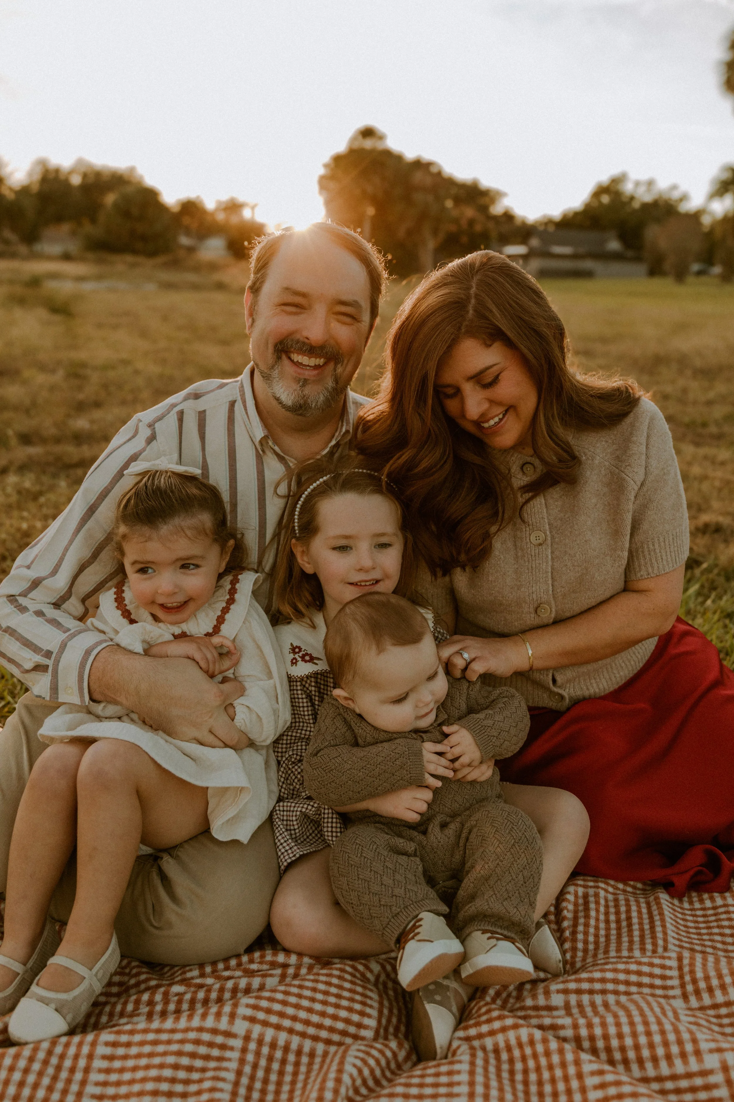 Family of five sitting together on a blanket at sunset, laughing and holding their young children during a relaxed outdoor photography session