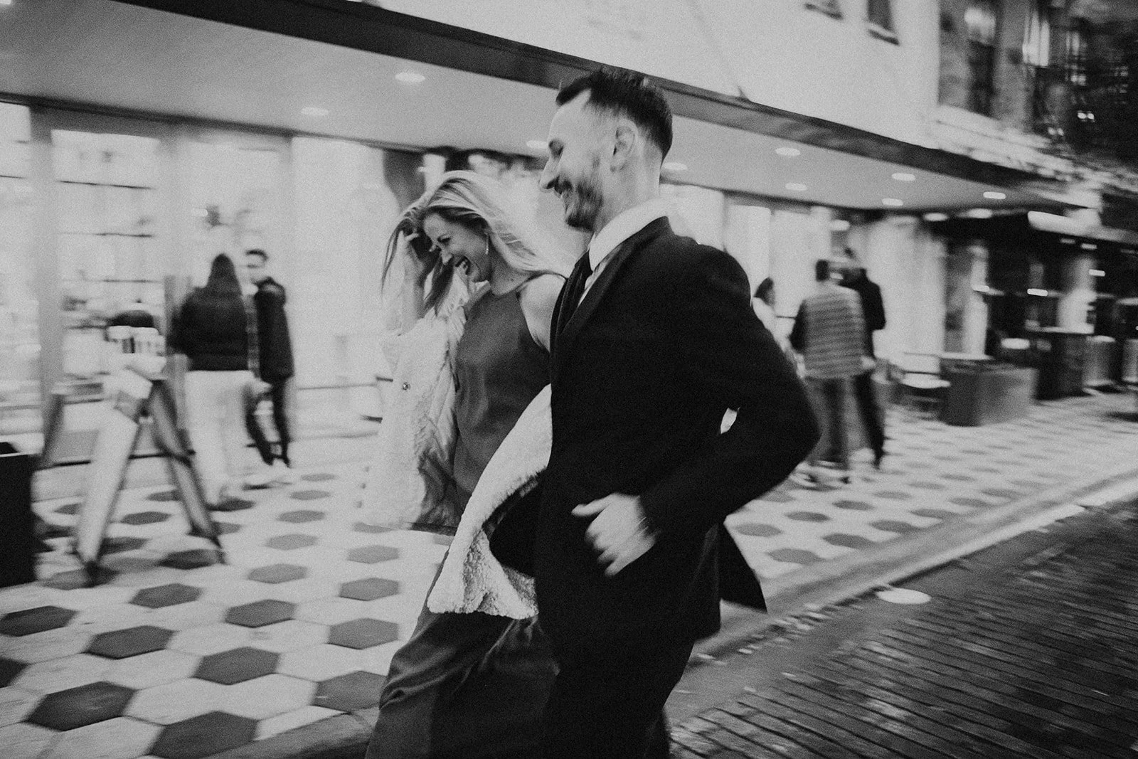 Black and white photo of a joyful couple walking down a city street at night. The woman is smiling and holding a coat, while the man, dressed in a suit, is also smiling. There are people and city buildings in the background.